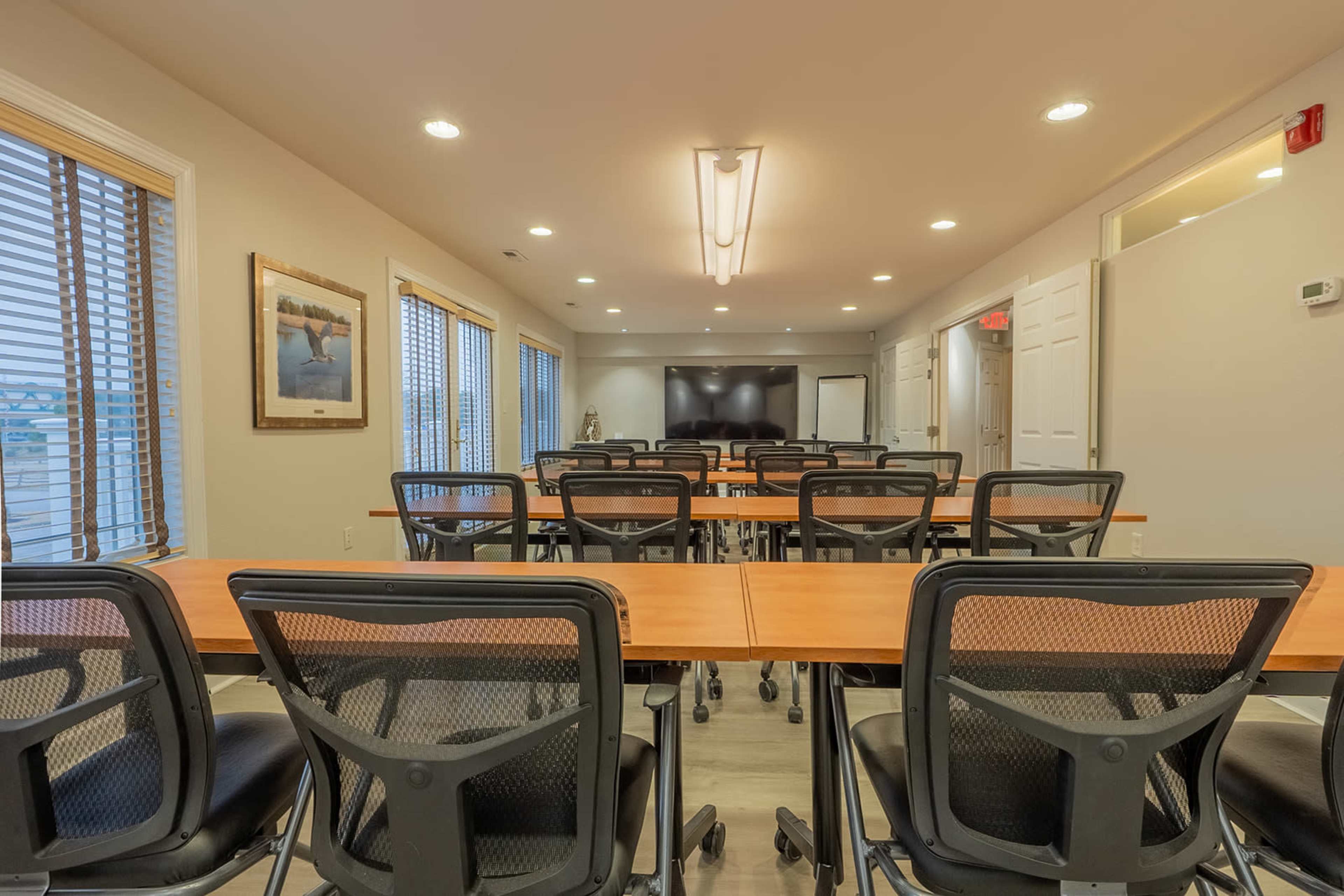 A conference room with rows of tables and chairs arranged for a meeting, featuring large windows and a screen at the front.