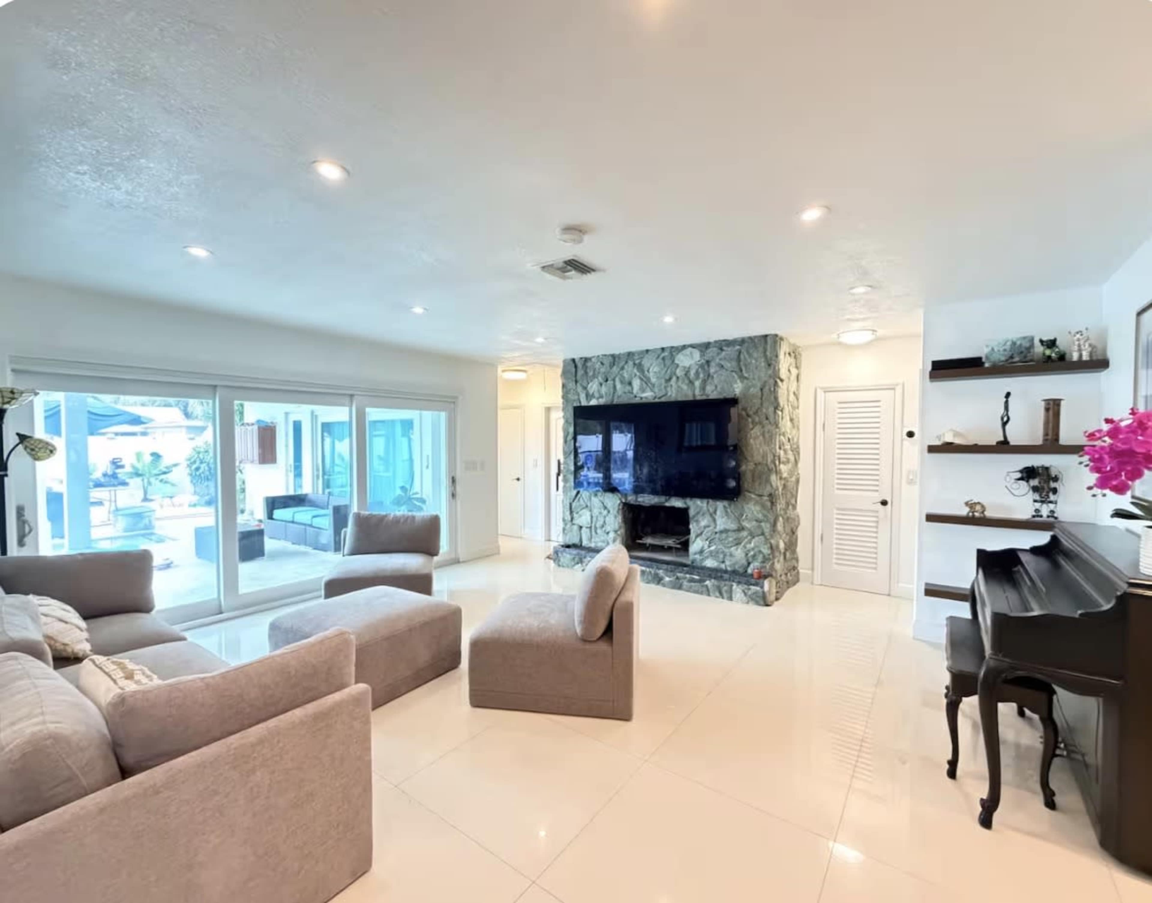 The living room features a light-colored sofa with matching armchairs, a stone fireplace, and a television mounted on the wall, surrounded by large windows that open to an outdoor space.