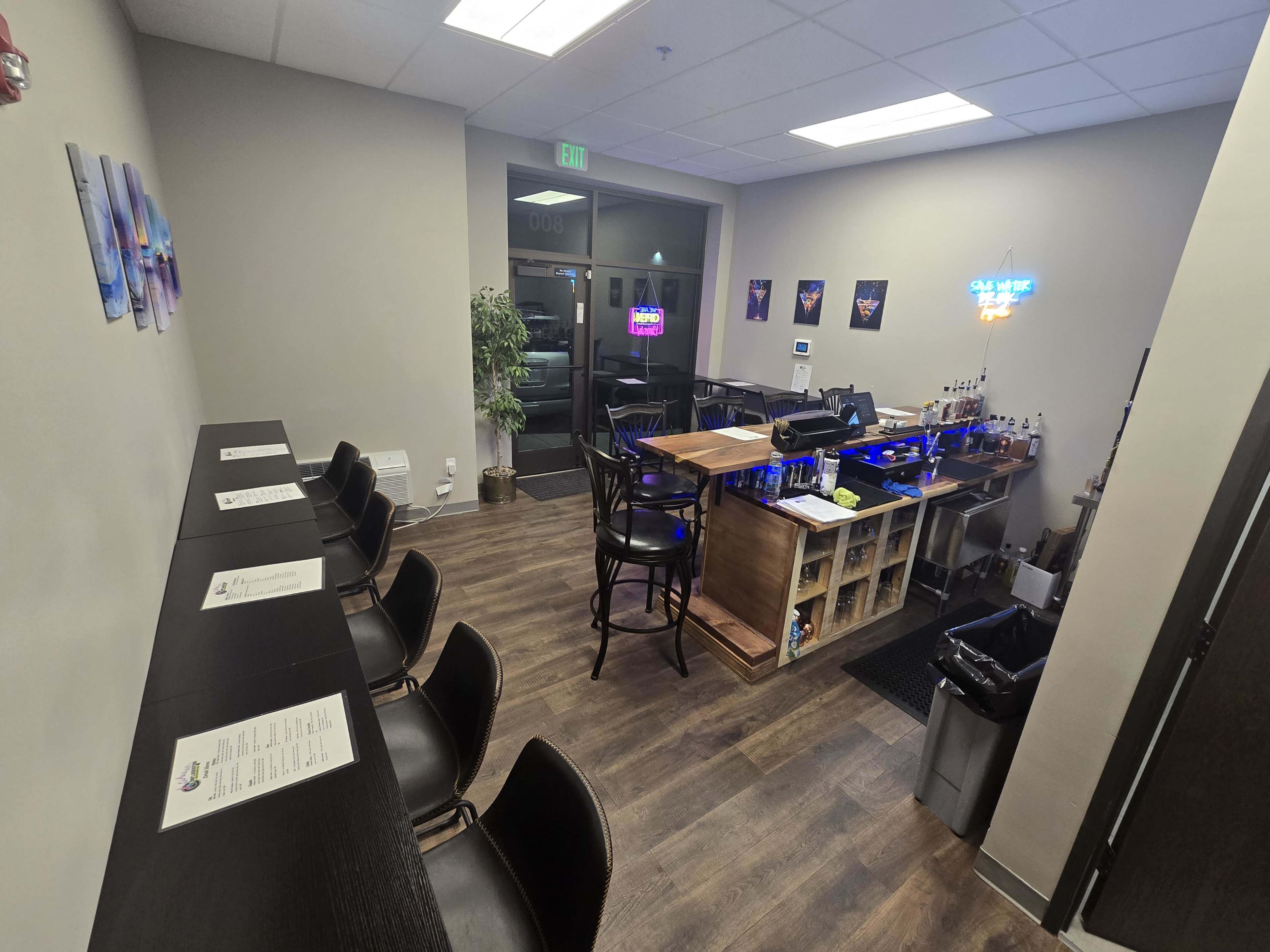 The image shows a small interior space featuring a bar with high stools, shelves of liquor, and a seating area with tables against the wall.