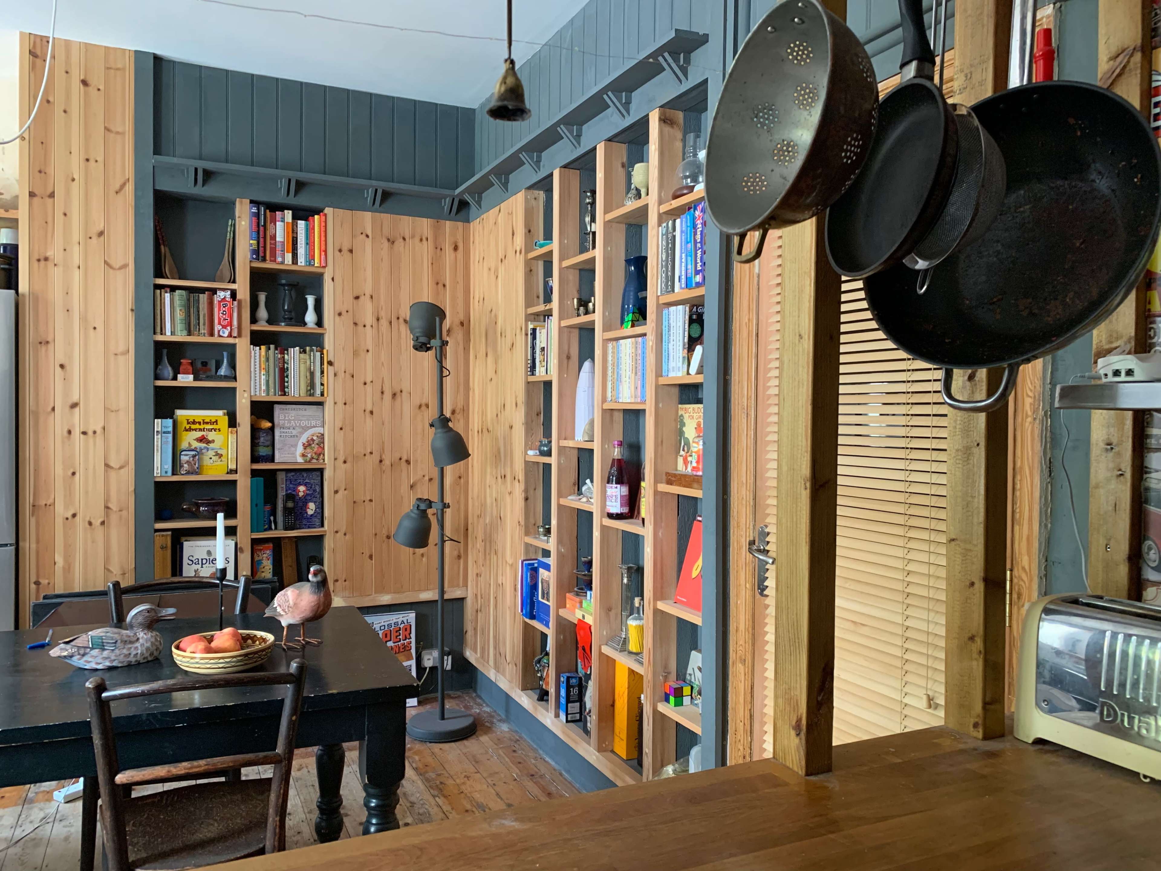 The image shows a cozy kitchen area with wooden walls, a black dining table, and shelves filled with books and various items.