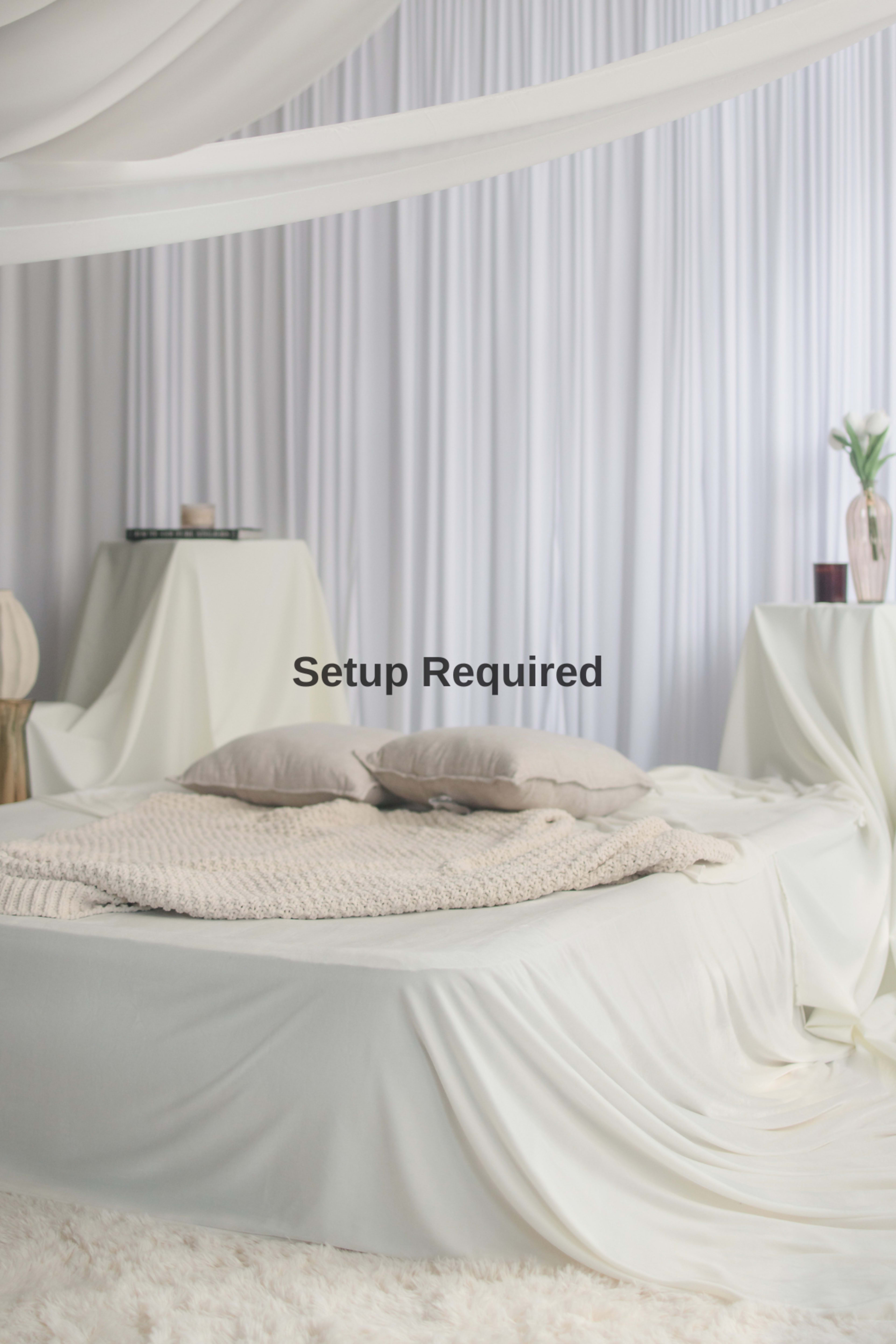 A neatly arranged bed with a beige blanket and two pillows is positioned in a softly lit room, surrounded by draped fabric and minimalistic decor.