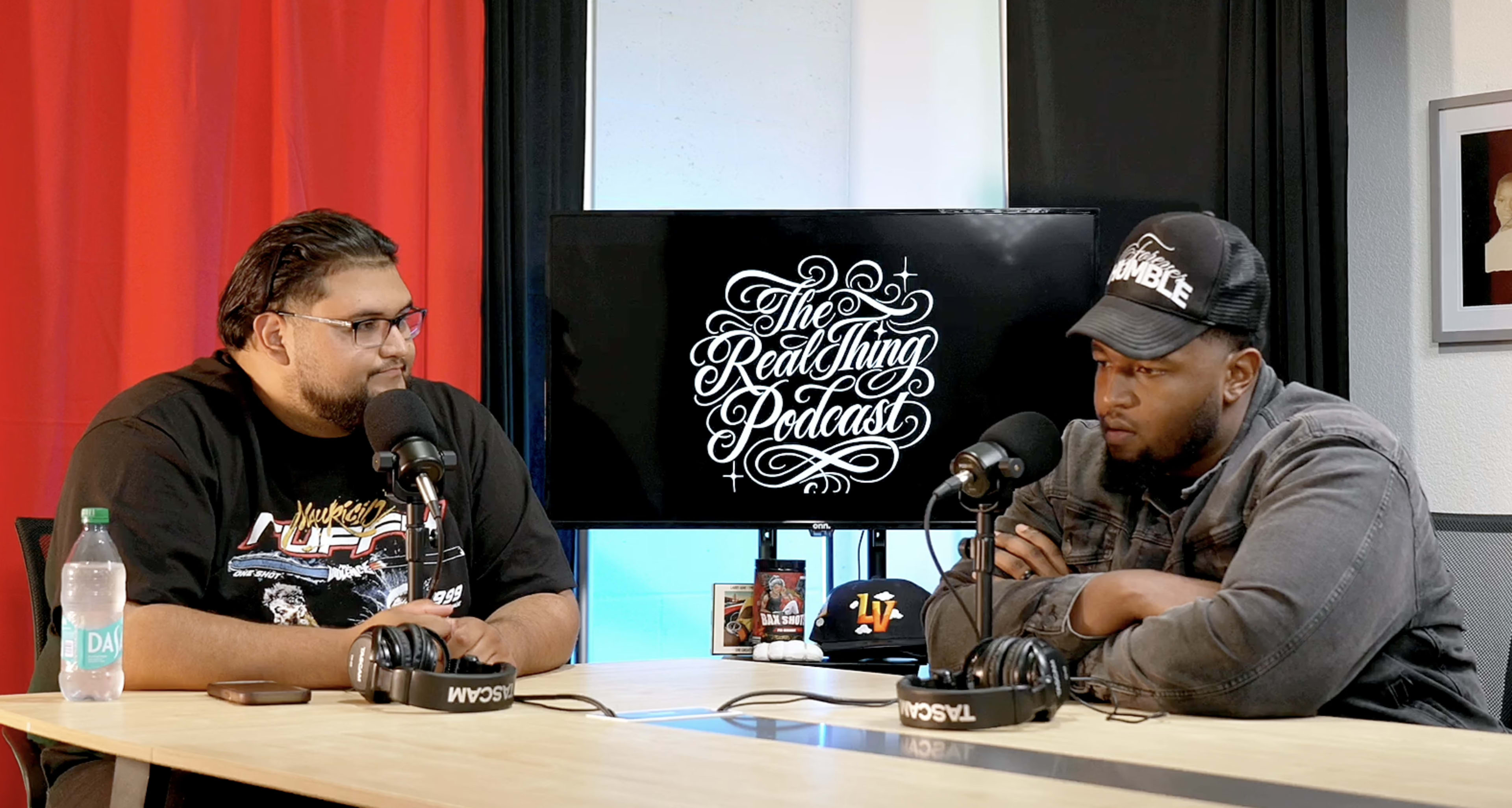 Two hosts sit at a table in a podcast studio, engaged in conversation with microphones in front of them, and a screen displaying "The Real Thing Podcast" logo in the background.
