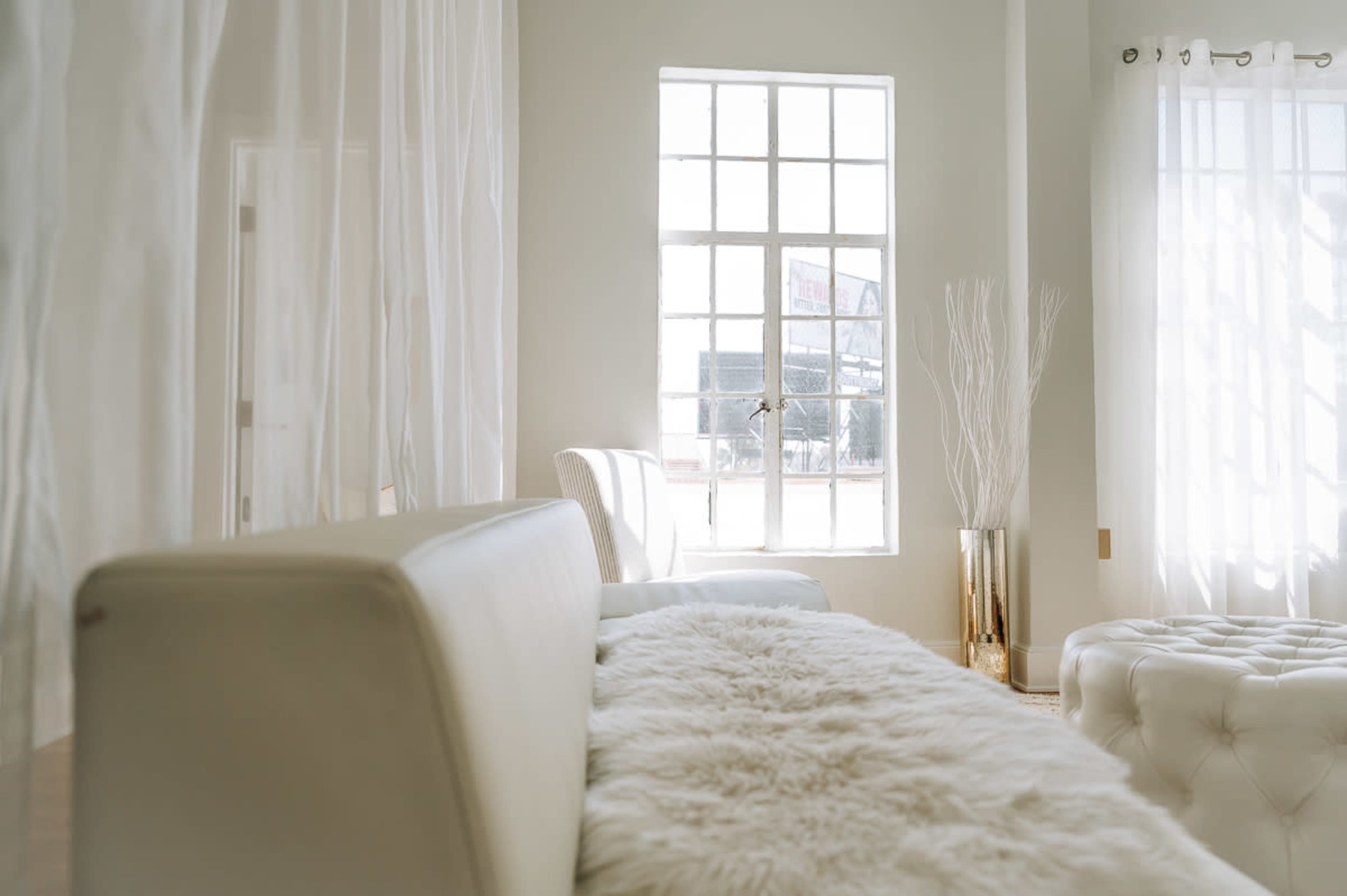A bright room features a white couch, a tufted ottoman, sheer curtains, and a tall vase with branches near a window.