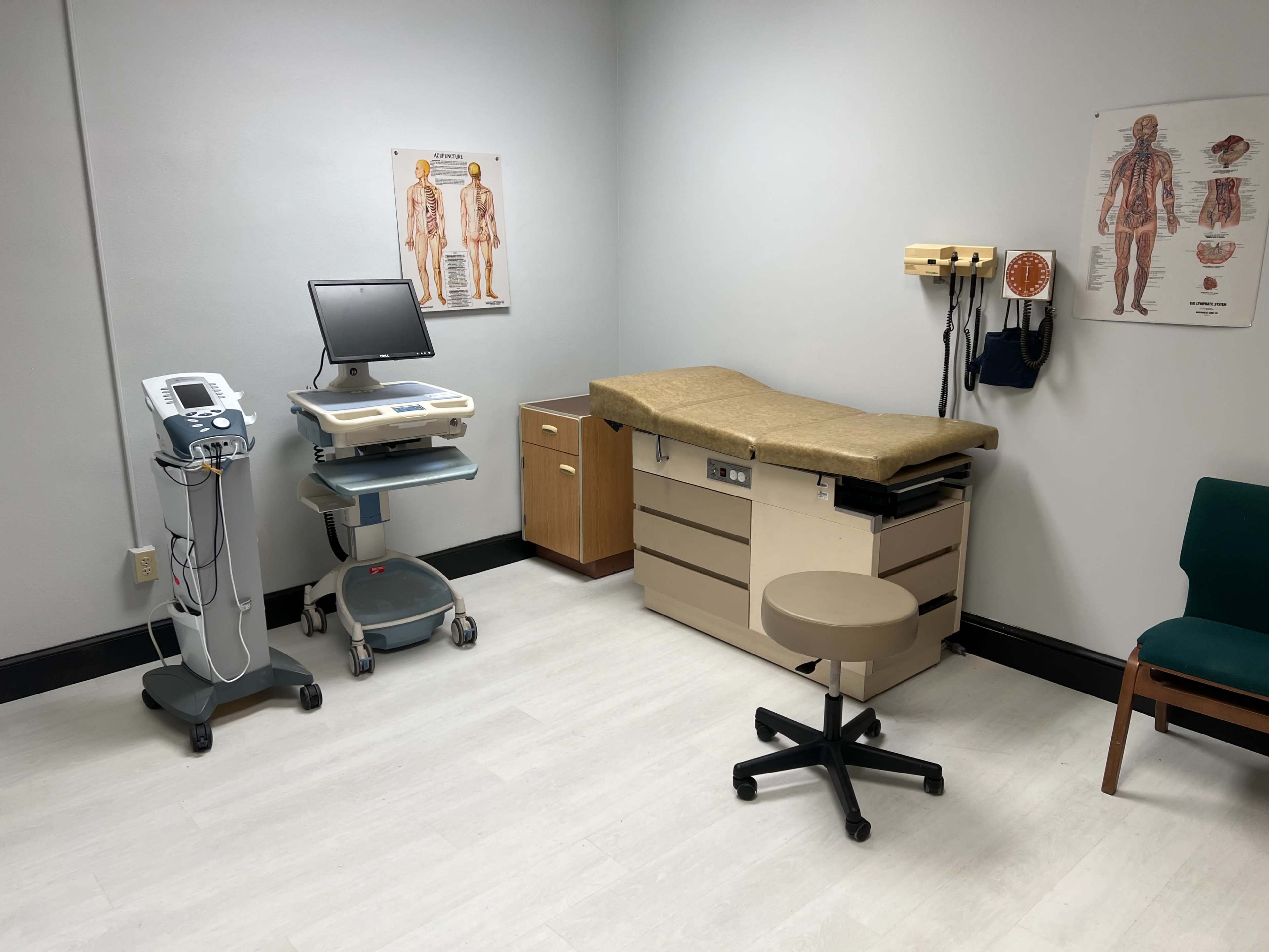 The image shows a medical examination room with a treatment table, medical equipment, a chair, and anatomical posters on the wall.
