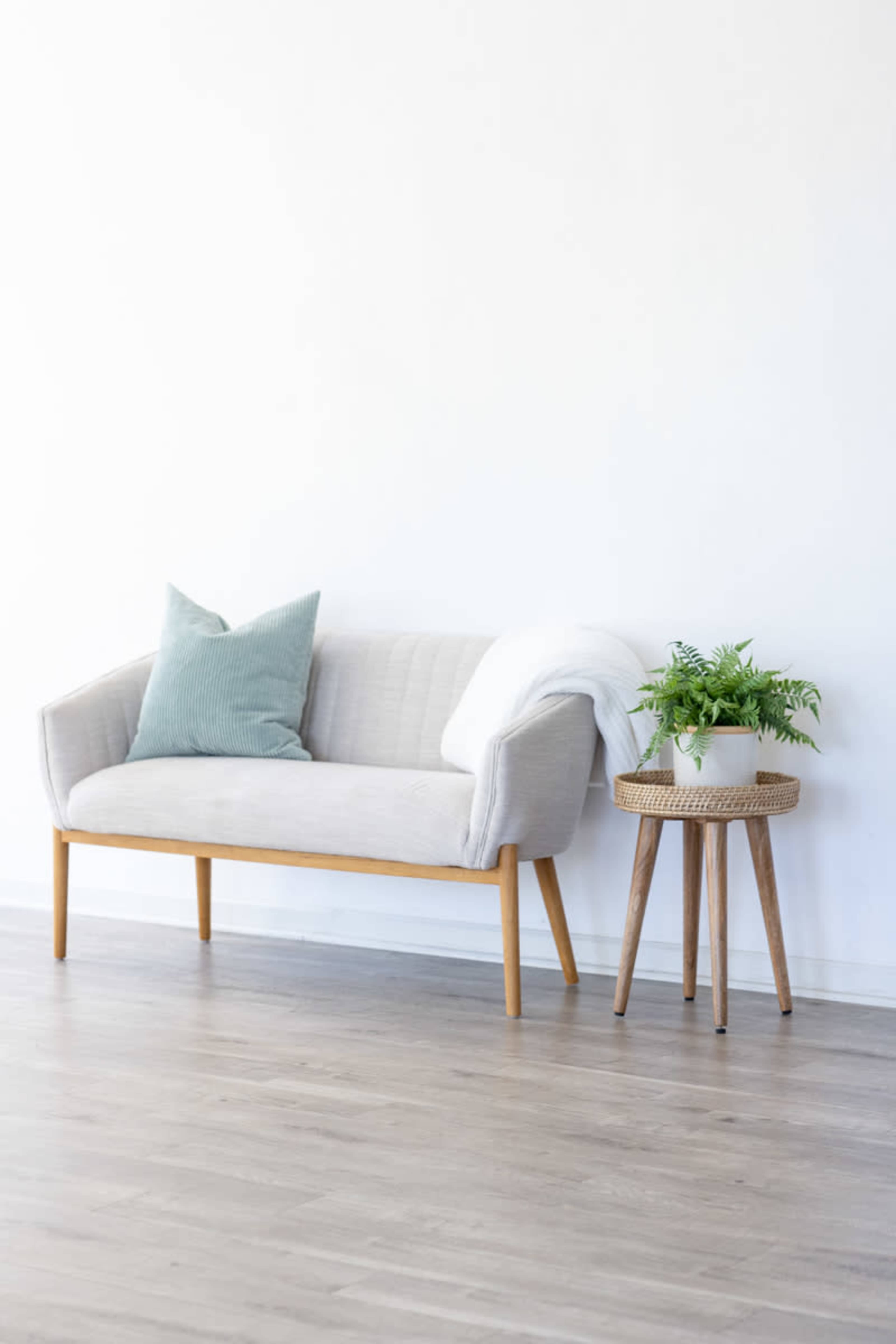 A light-colored sofa with a blue-green pillow sits beside a small round table with a potted plant on it against a white wall.
