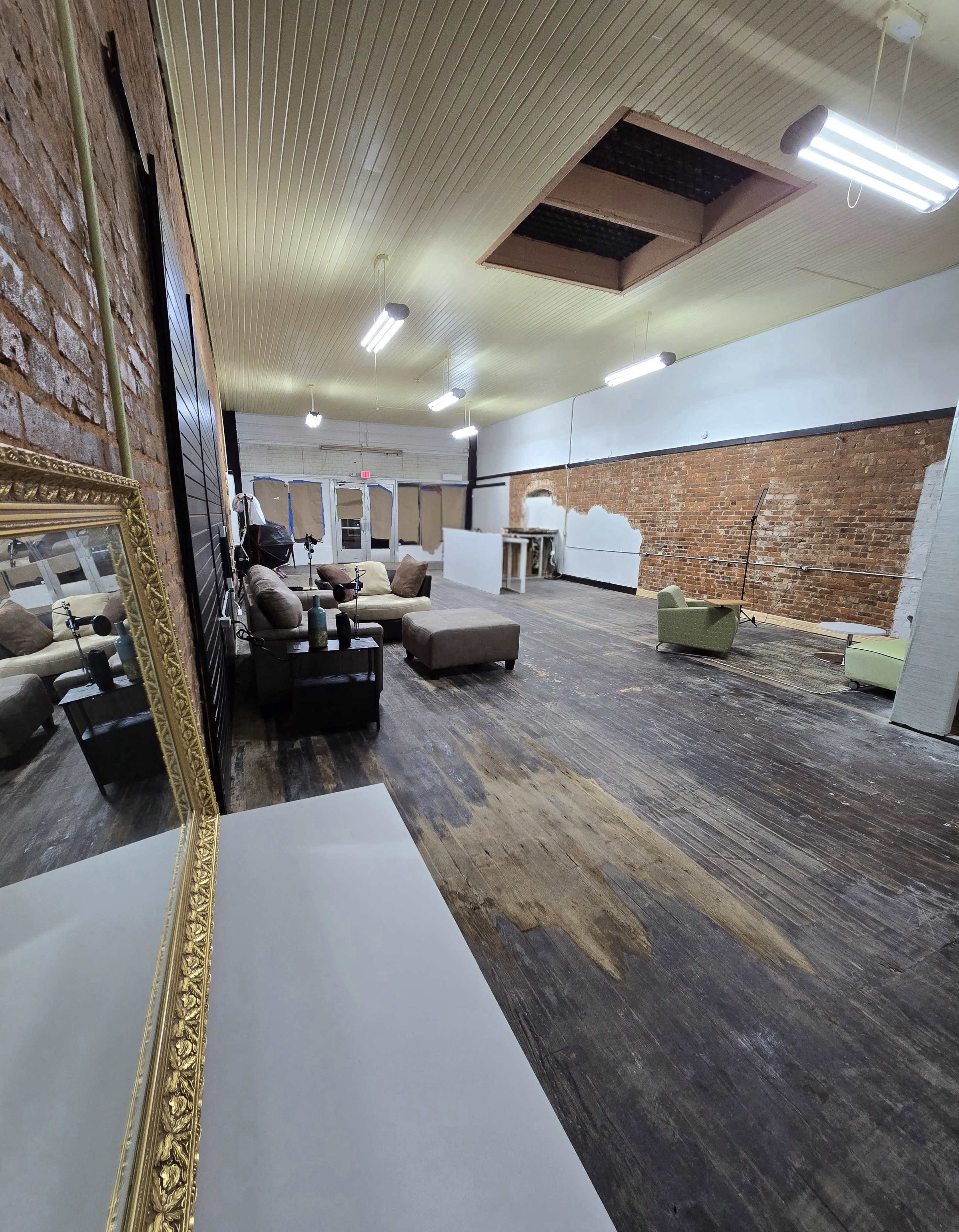 The image shows an empty interior space with exposed brick walls, wooden flooring, and various furniture pieces scattered throughout.