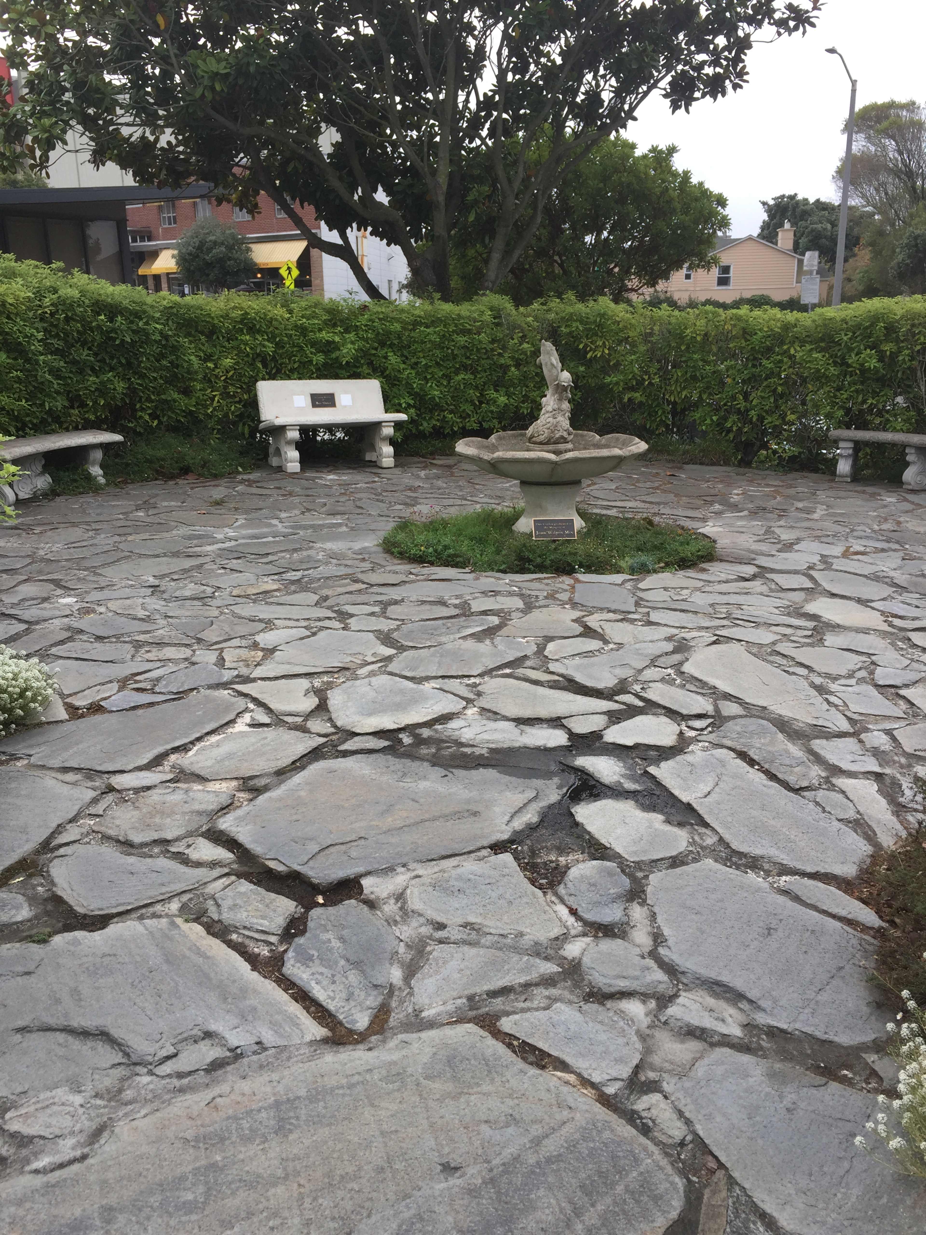 A circular stone courtyard with a fountain at its center, surrounded by benches and greenery.
