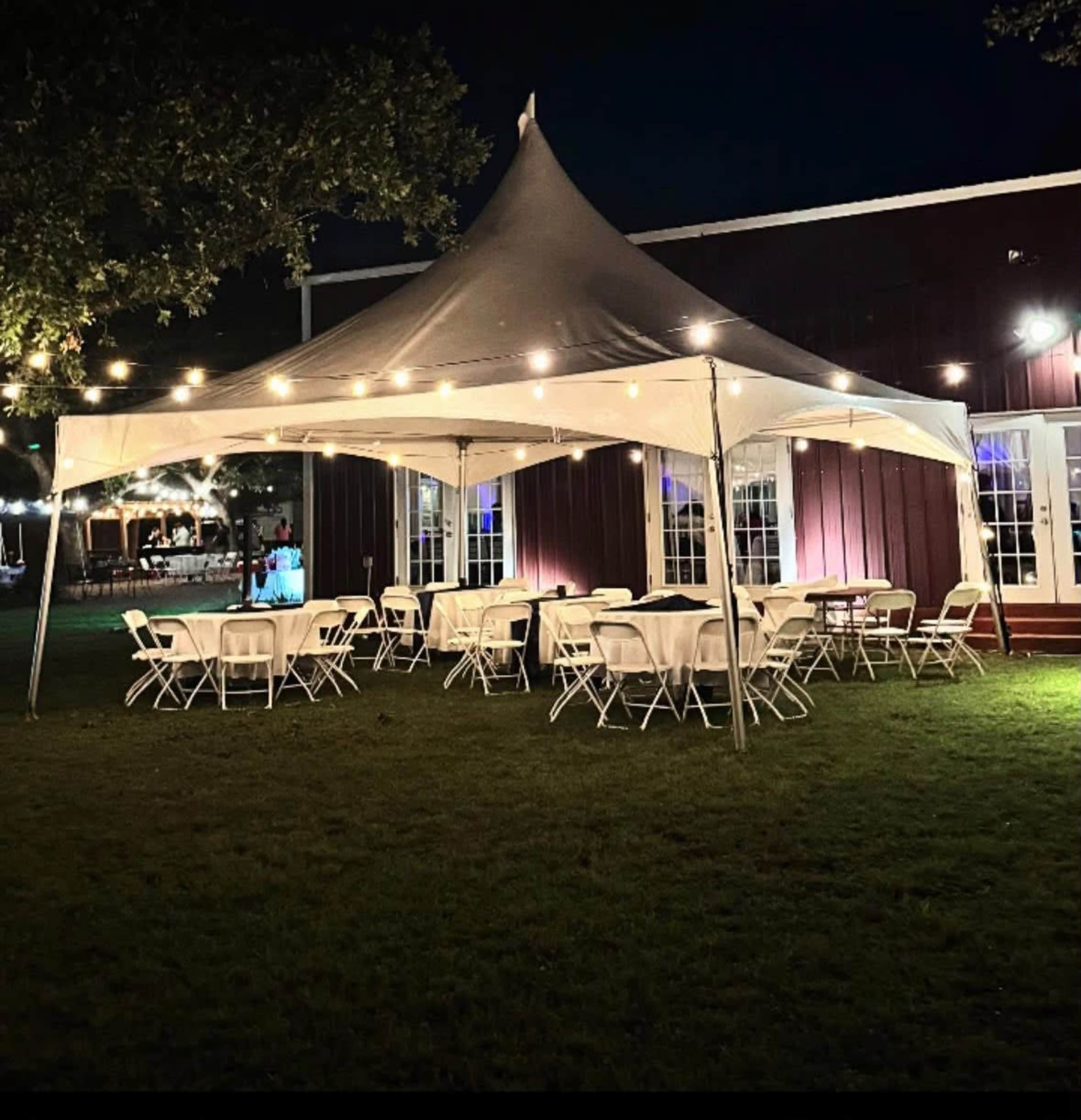 A large white tent with several round tables and chairs is set up on a grassy area next to a building, illuminated by string lights at night.