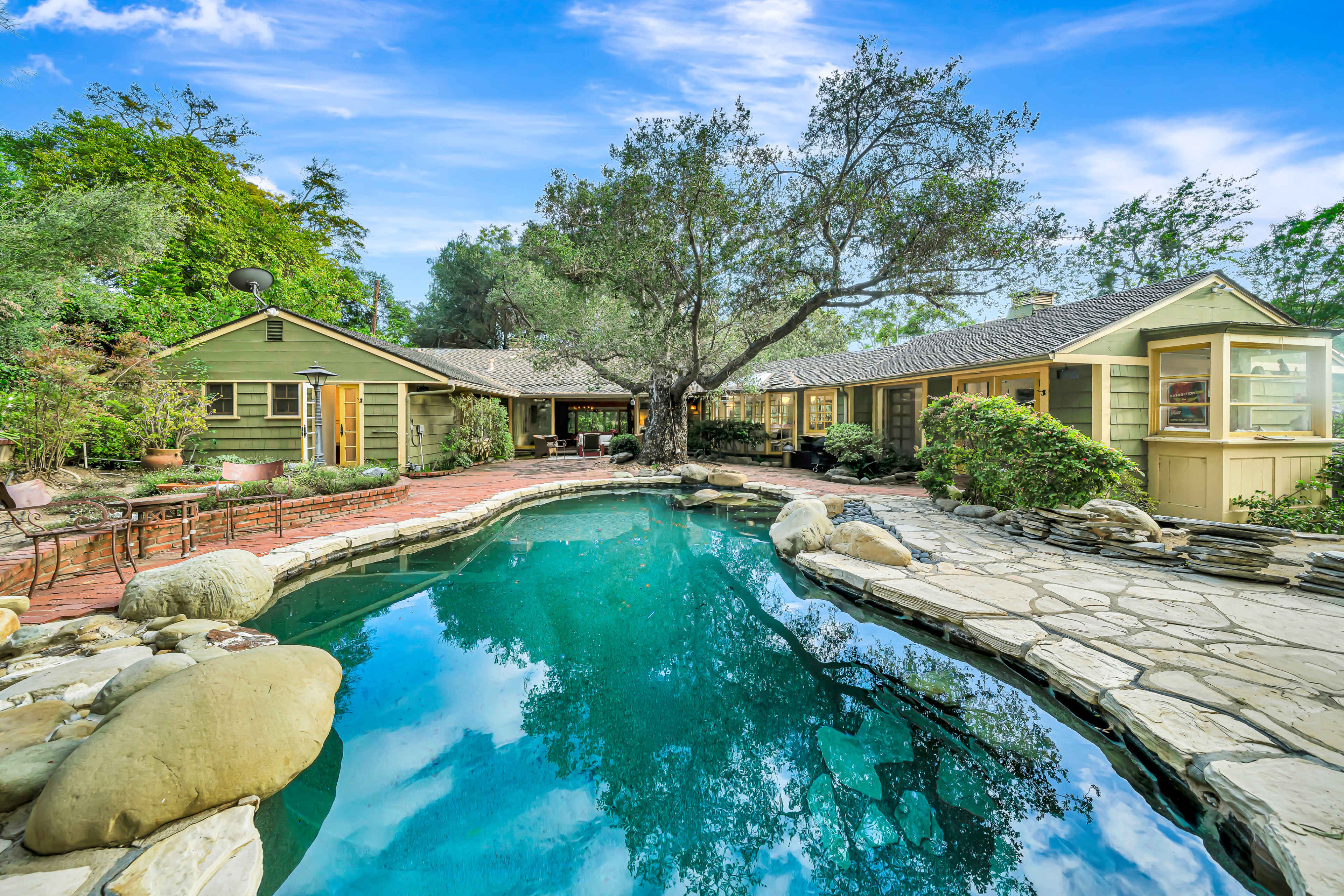 The image shows a ranch-style house with a pool surrounded by a stone patio and lush greenery.