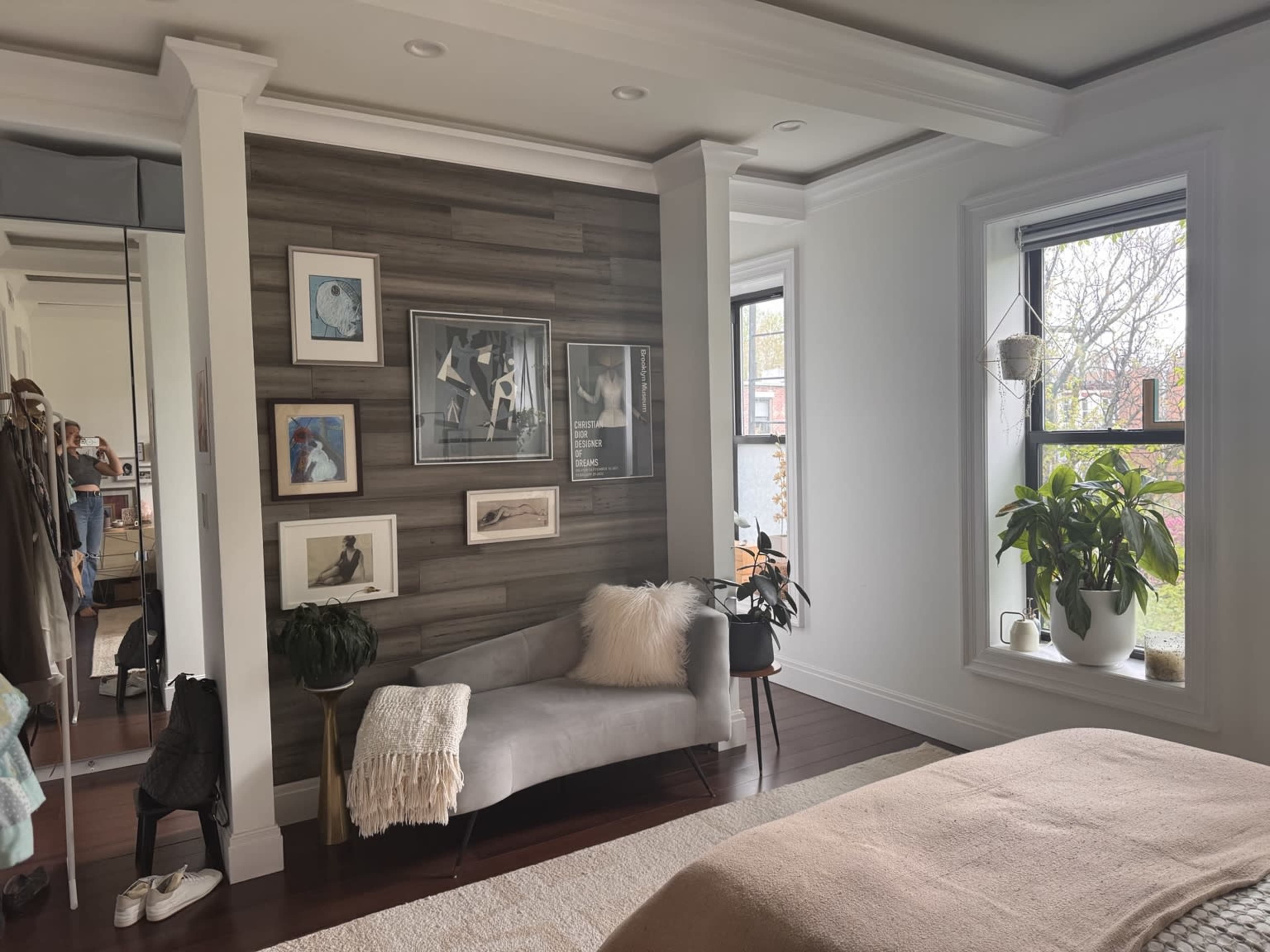 The image shows a modern bedroom featuring a gray sofa against a wood-paneled wall adorned with framed artwork, along with a window that allows natural light and a potted plant nearby.