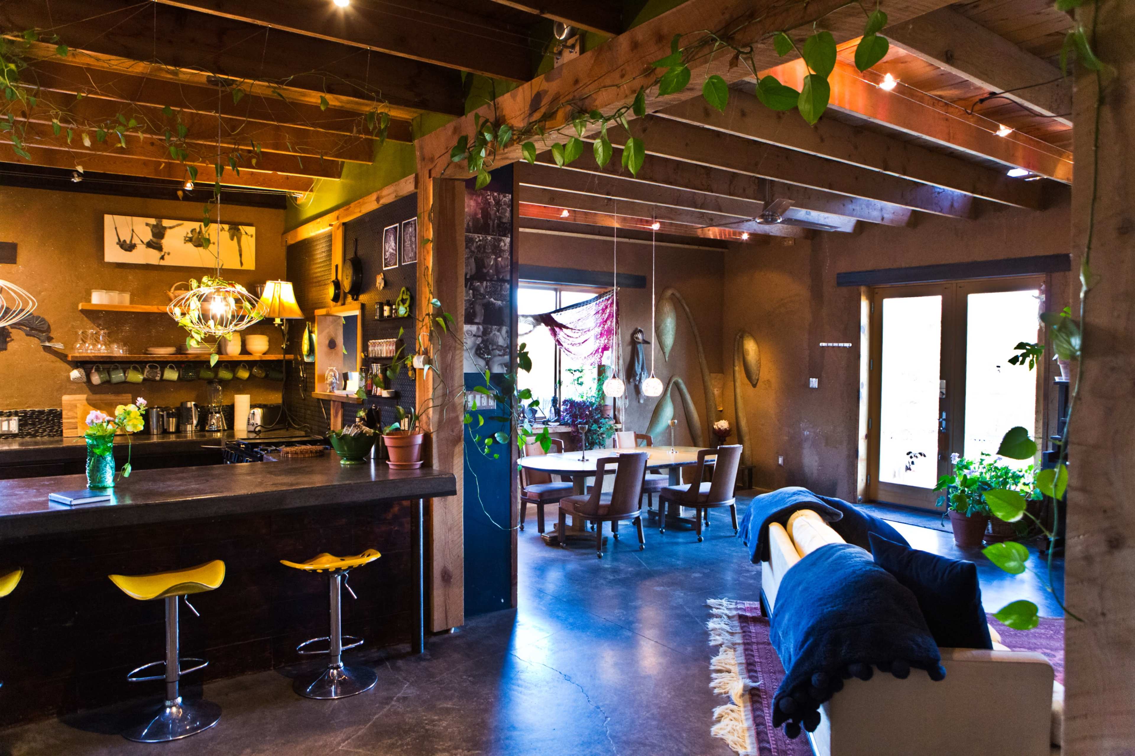 The image shows a modern, open-concept living space featuring a kitchen bar with yellow stools, a dining area, and various plants throughout.