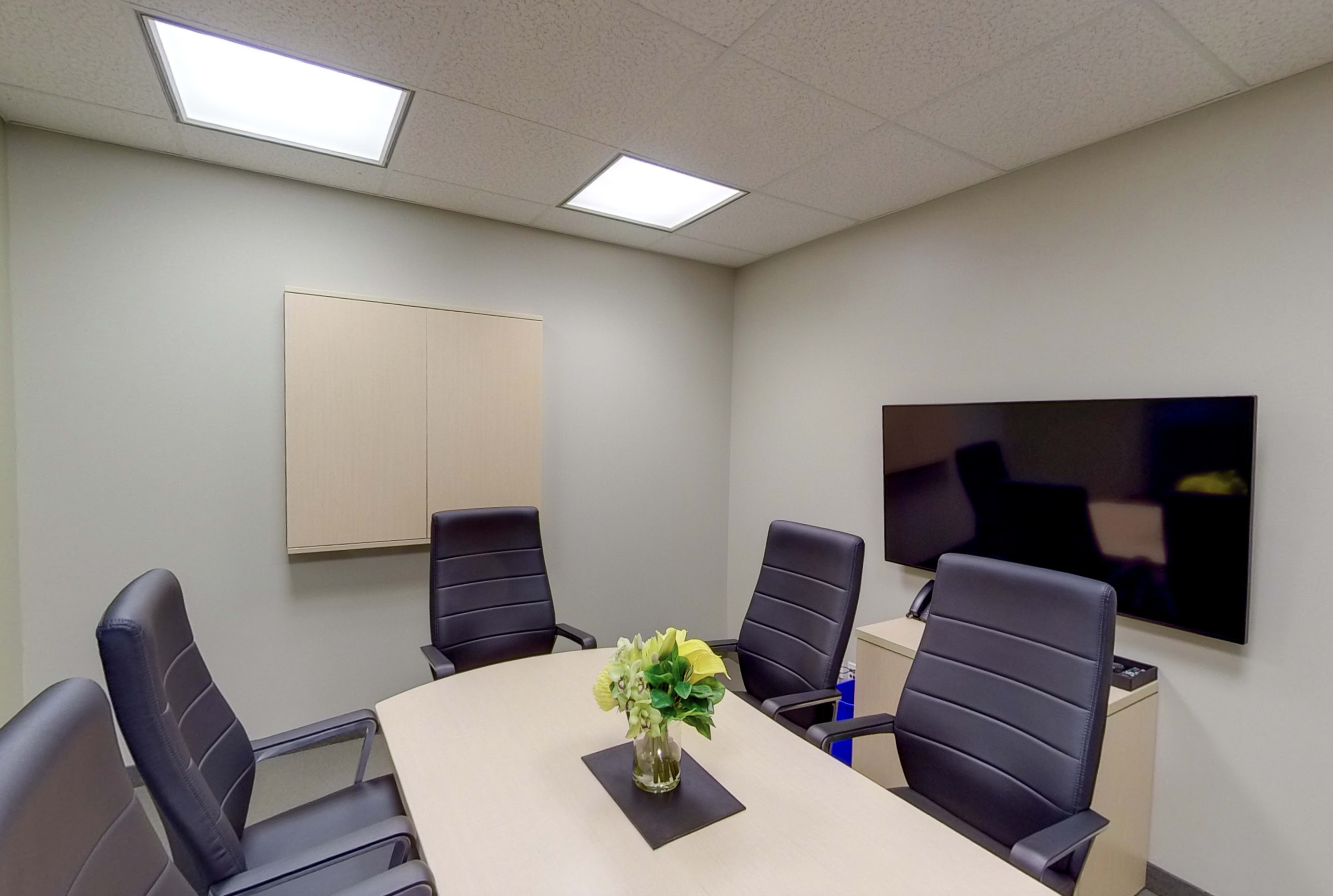 A conference room features a rectangular table surrounded by four black leather chairs, with a small vase of flowers in the center and a wall-mounted TV on one side.