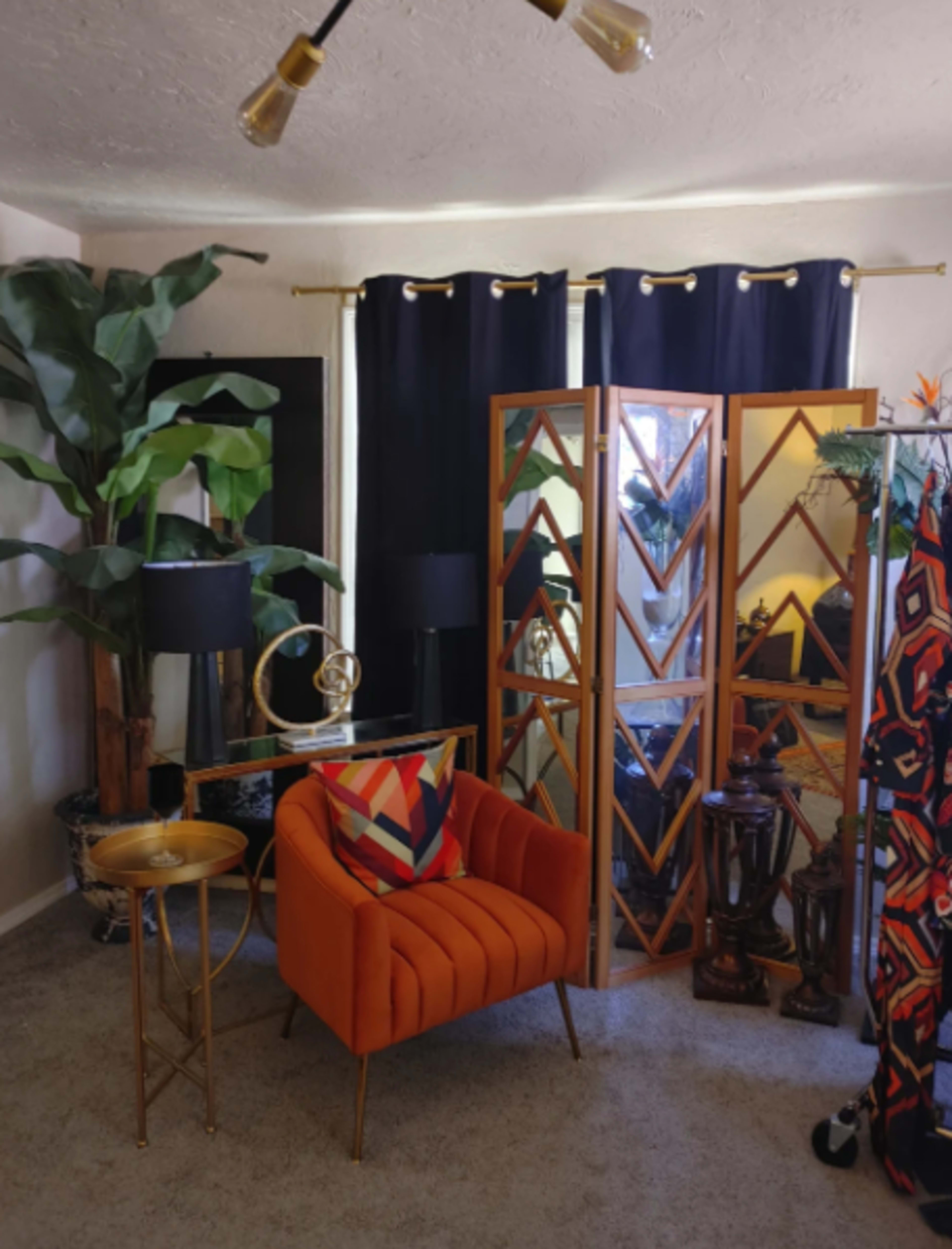 A corner of a room features an orange armchair, a mirrored screen, potted plants, and decorative lamps, surrounded by dark curtains.