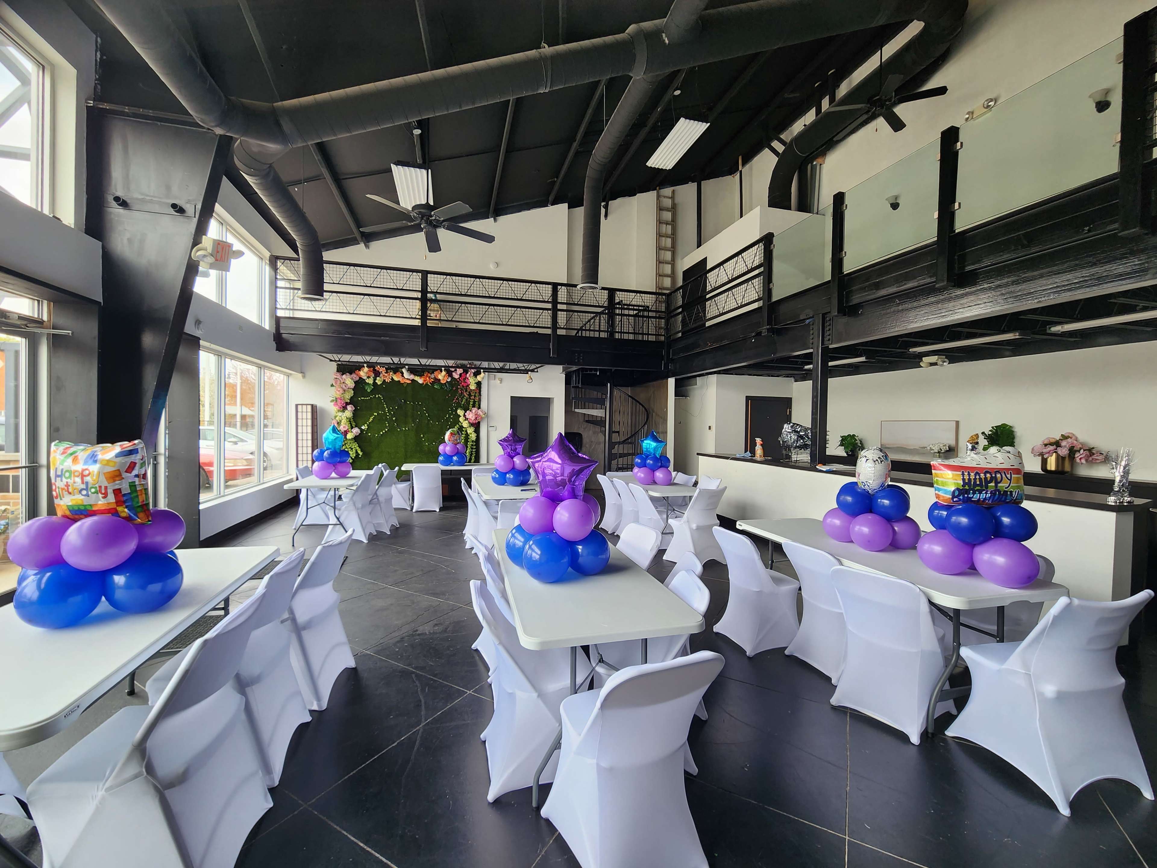 A decorated event space with white tables covered in colorful balloon centerpieces and a vibrant floral backdrop.