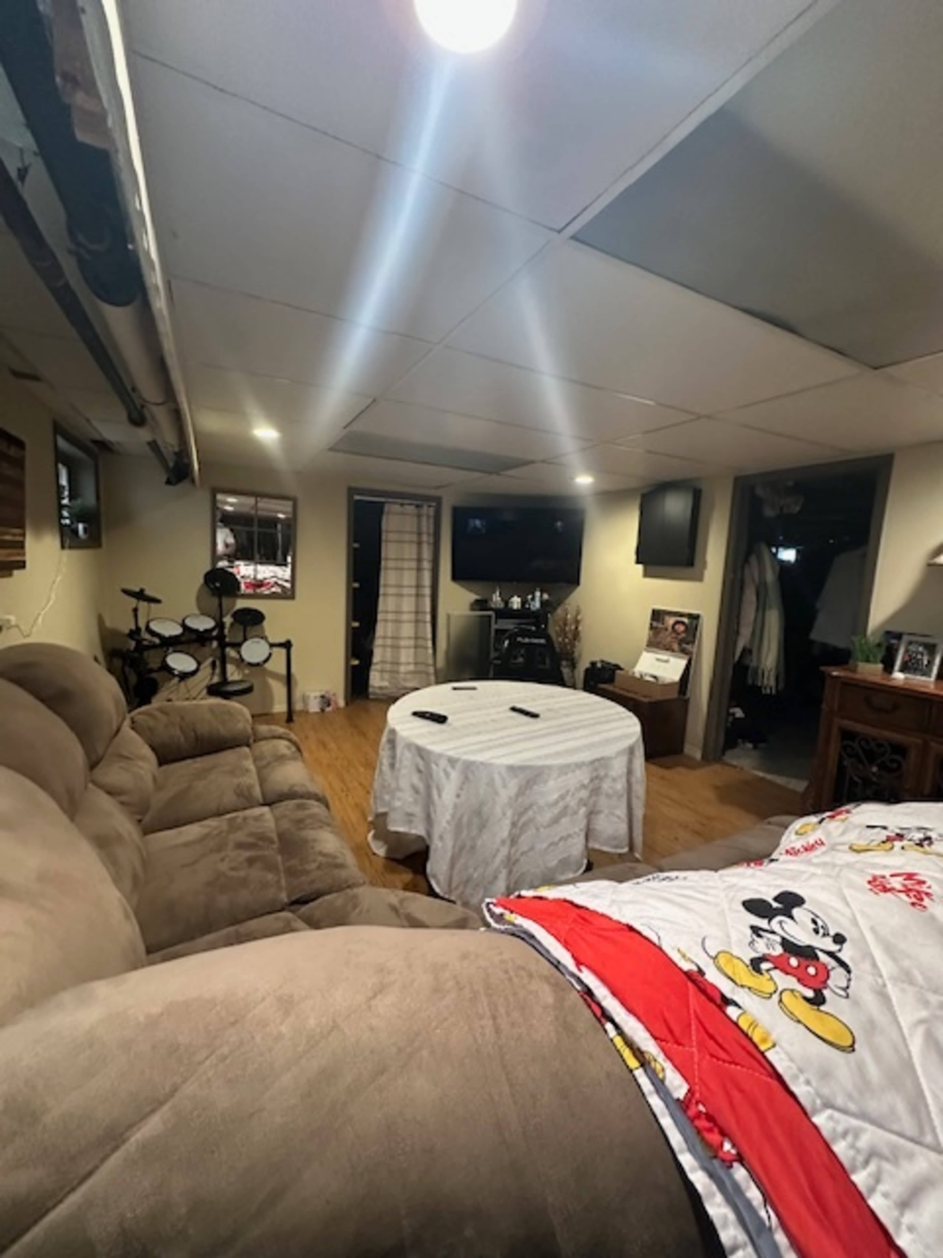 The image shows a basement living area with a sectional couch, a round table covered with a white tablecloth, and a TV mounted on the wall.