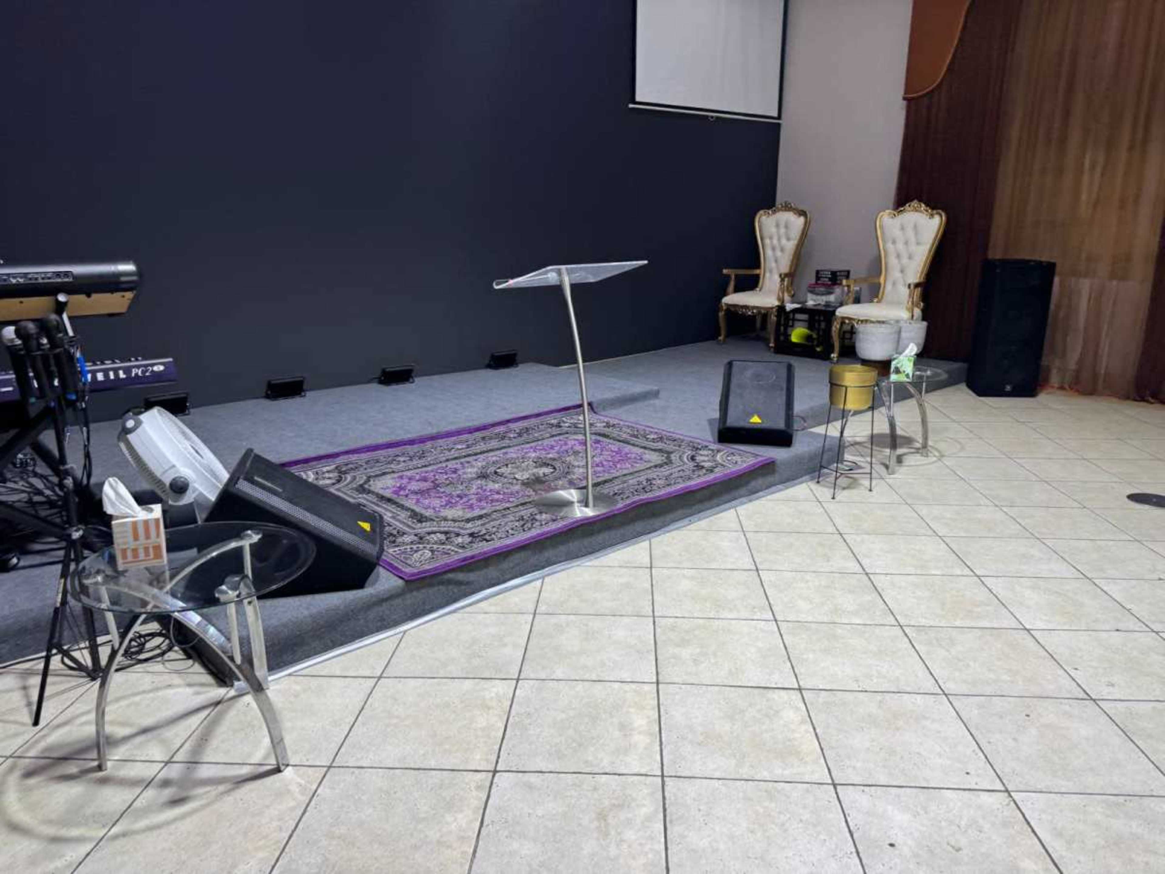 The scene shows a stage area in a venue, featuring a clear podium on a decorative carpet, flanked by two ornate chairs and various audio equipment.