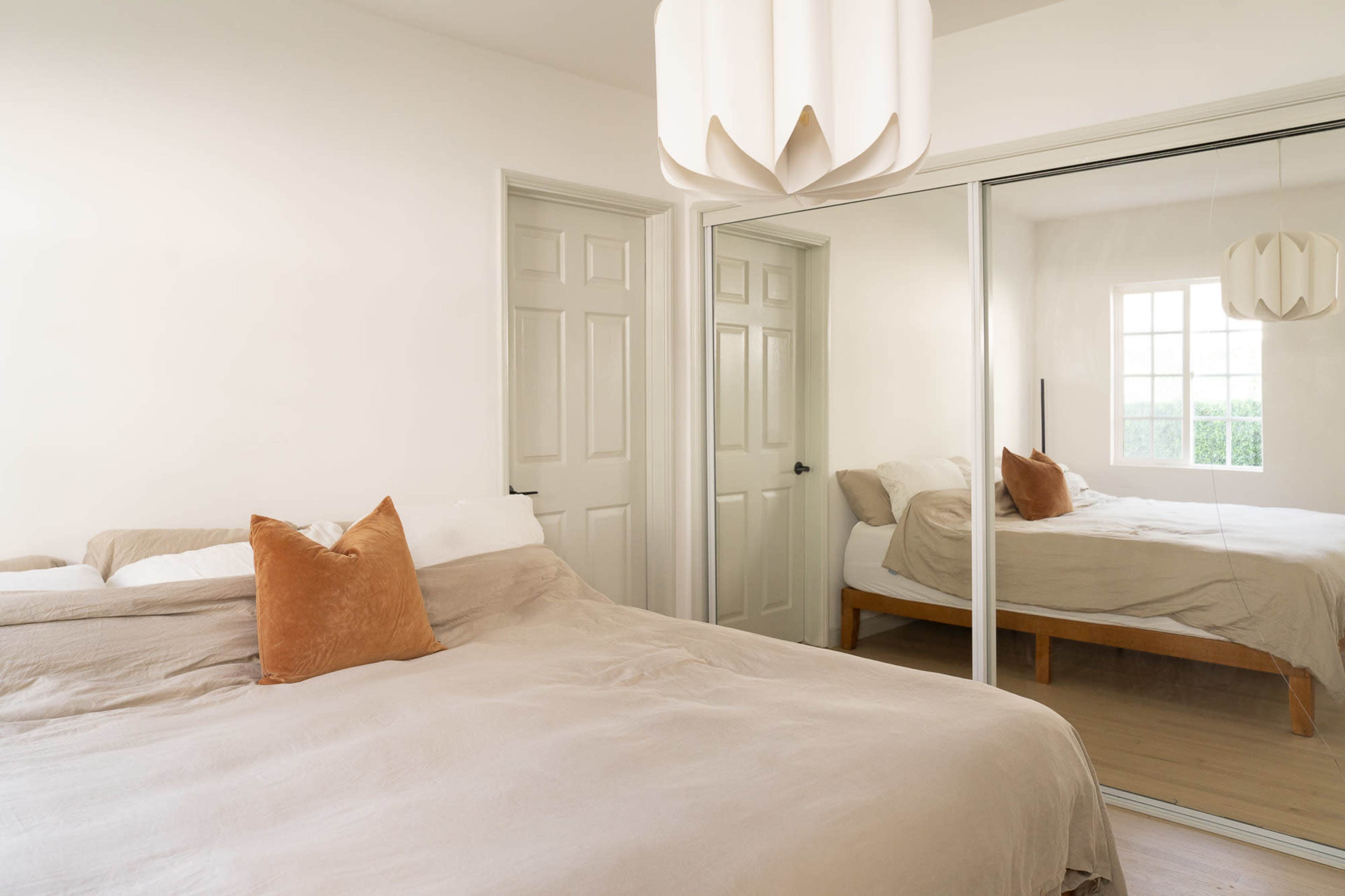 A minimalist bedroom features a light-colored bed with a decorative pillow, sliding mirrored closet doors, and a pendant light fixture.