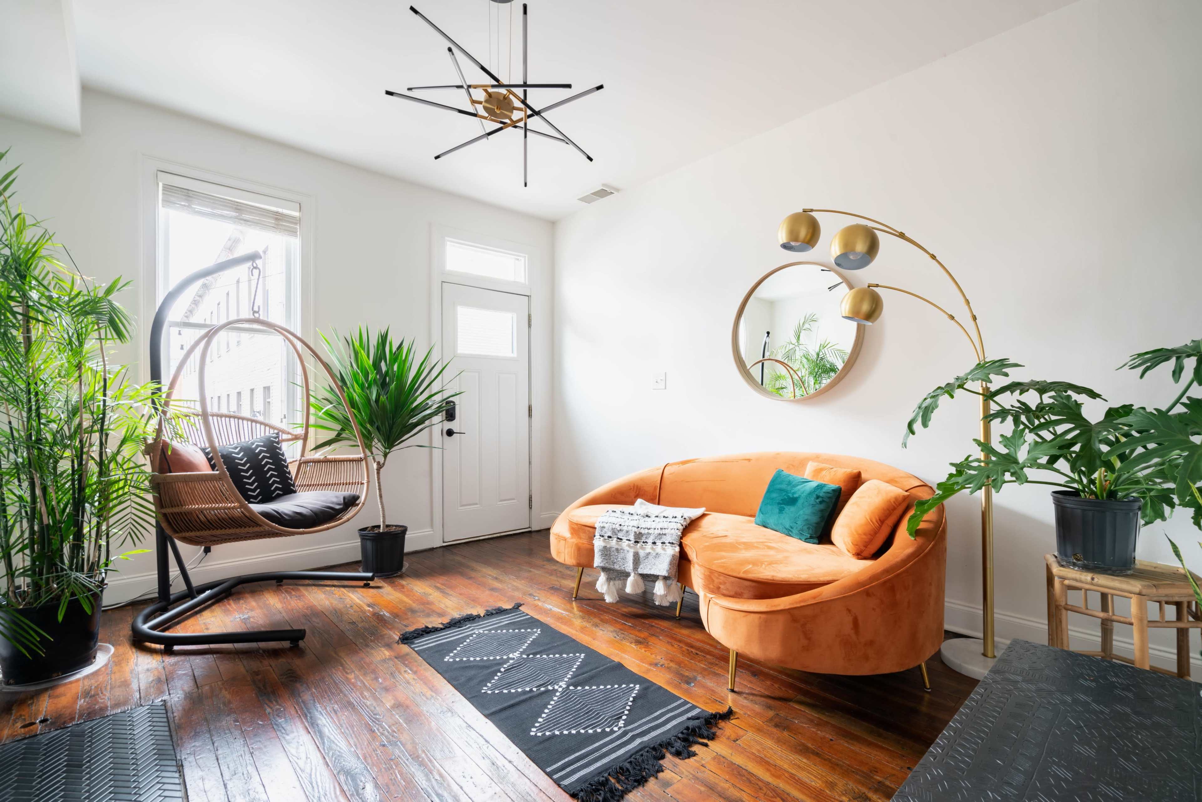 A modern living space featuring an orange curved sofa, a hanging chair, various indoor plants, and a round mirror on a white wall.