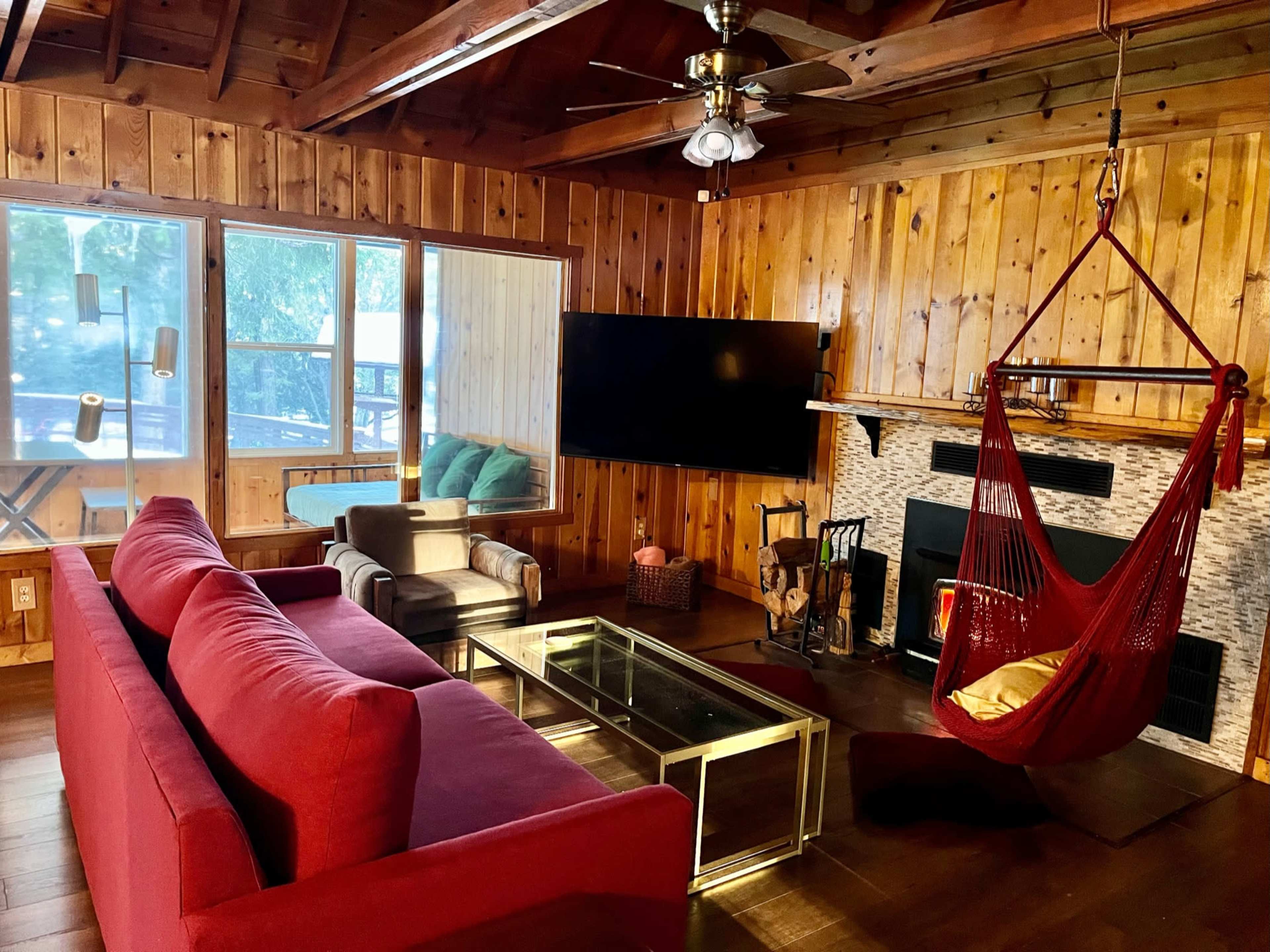 A cozy living room featuring wooden walls, a red sofa, a glass coffee table, and a hanging chair beside a fireplace.