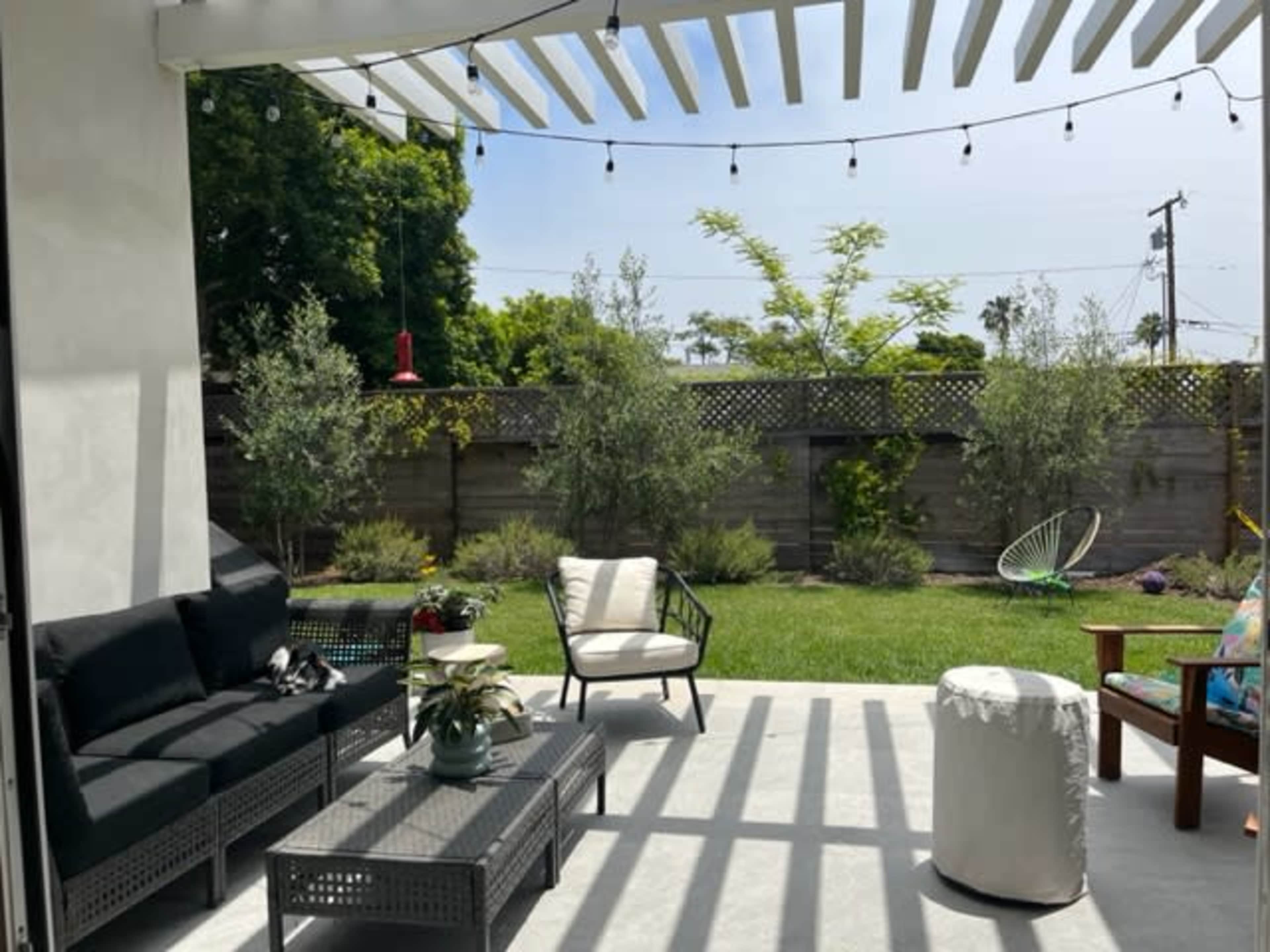 A patio area features a seating arrangement with black furniture, a small table, and a white ottoman, surrounded by greenery and a lawn under a pergola with hanging lights.