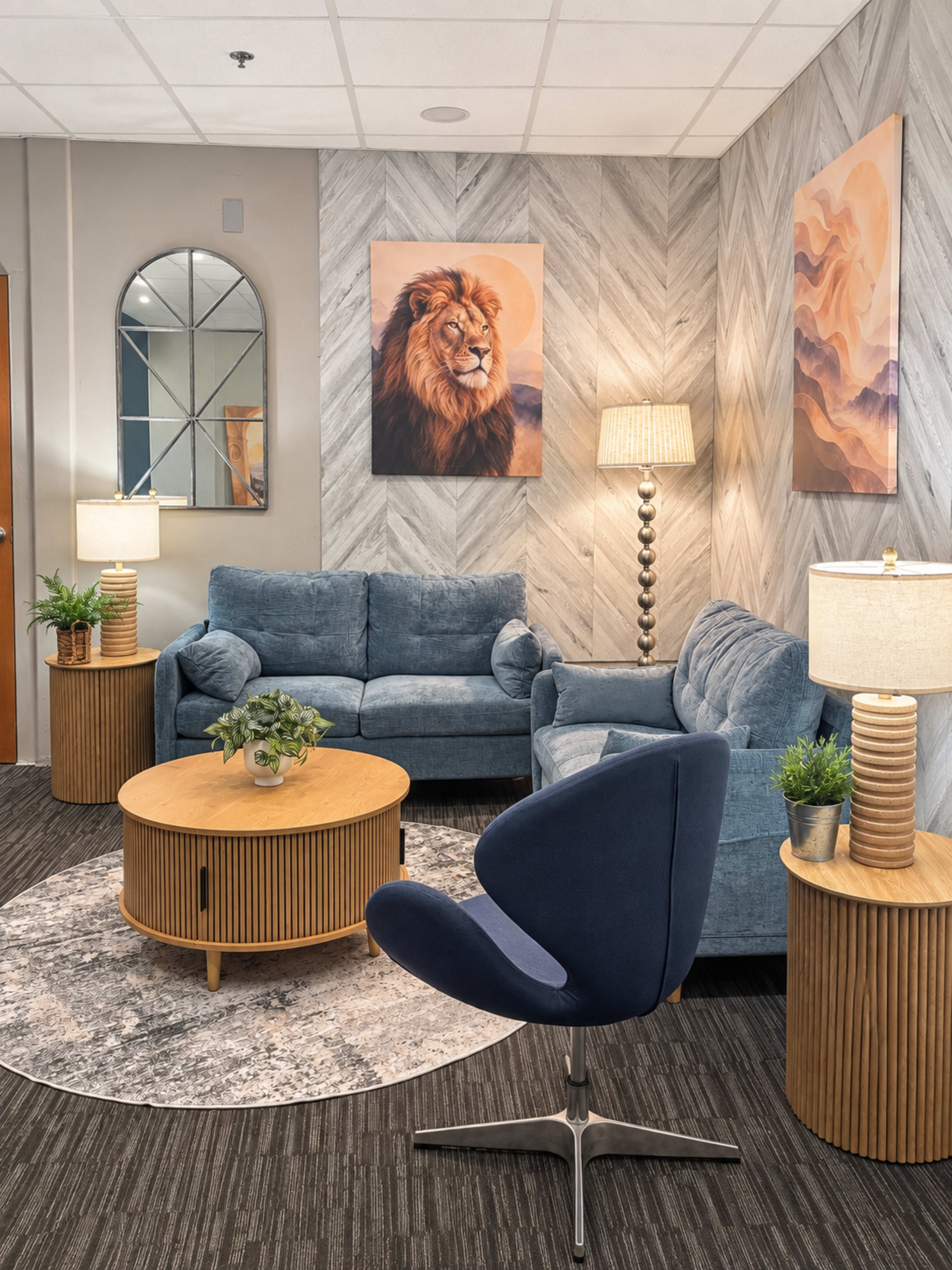 The image shows a cozy lounge area featuring blue upholstered sofas, a round wooden coffee table, two side tables with lamps, and wall art depicting a lion and abstract designs.
