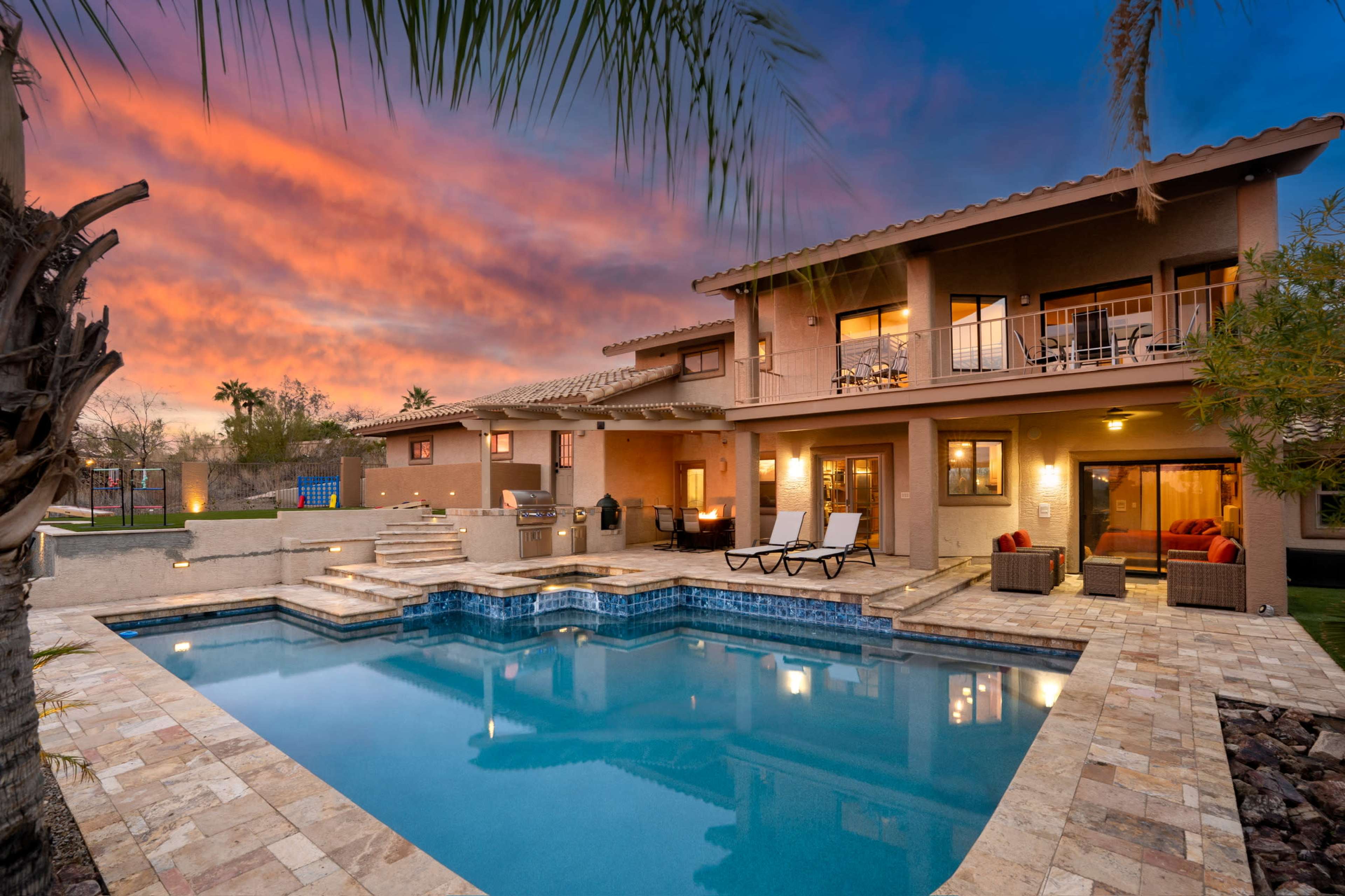 A two-story house with a pool is set against a colorful sunset, featuring outdoor seating areas and palm trees.