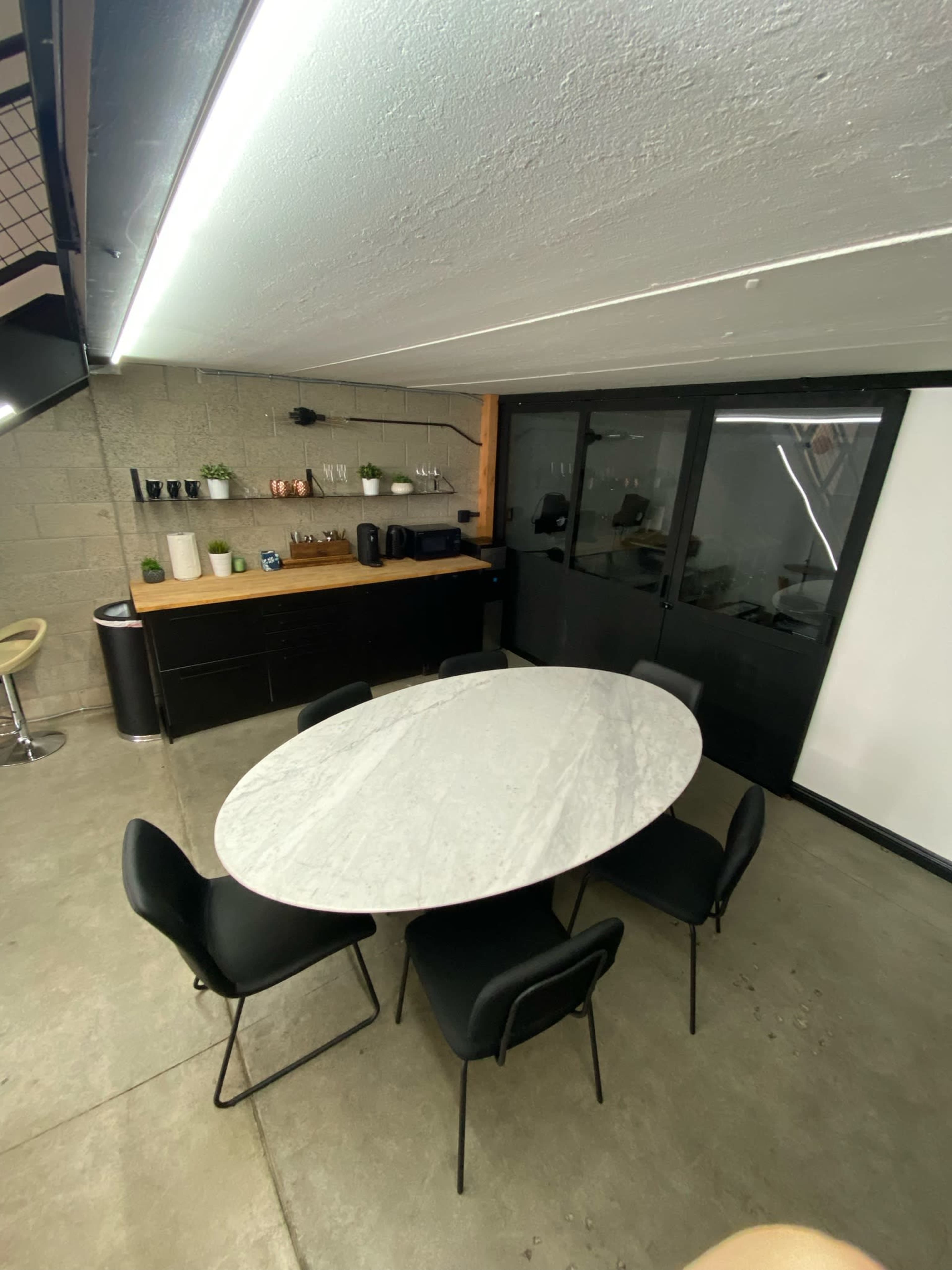 A spacious meeting room features a marble oval table surrounded by six black chairs, with a kitchen area and cabinets in the background.