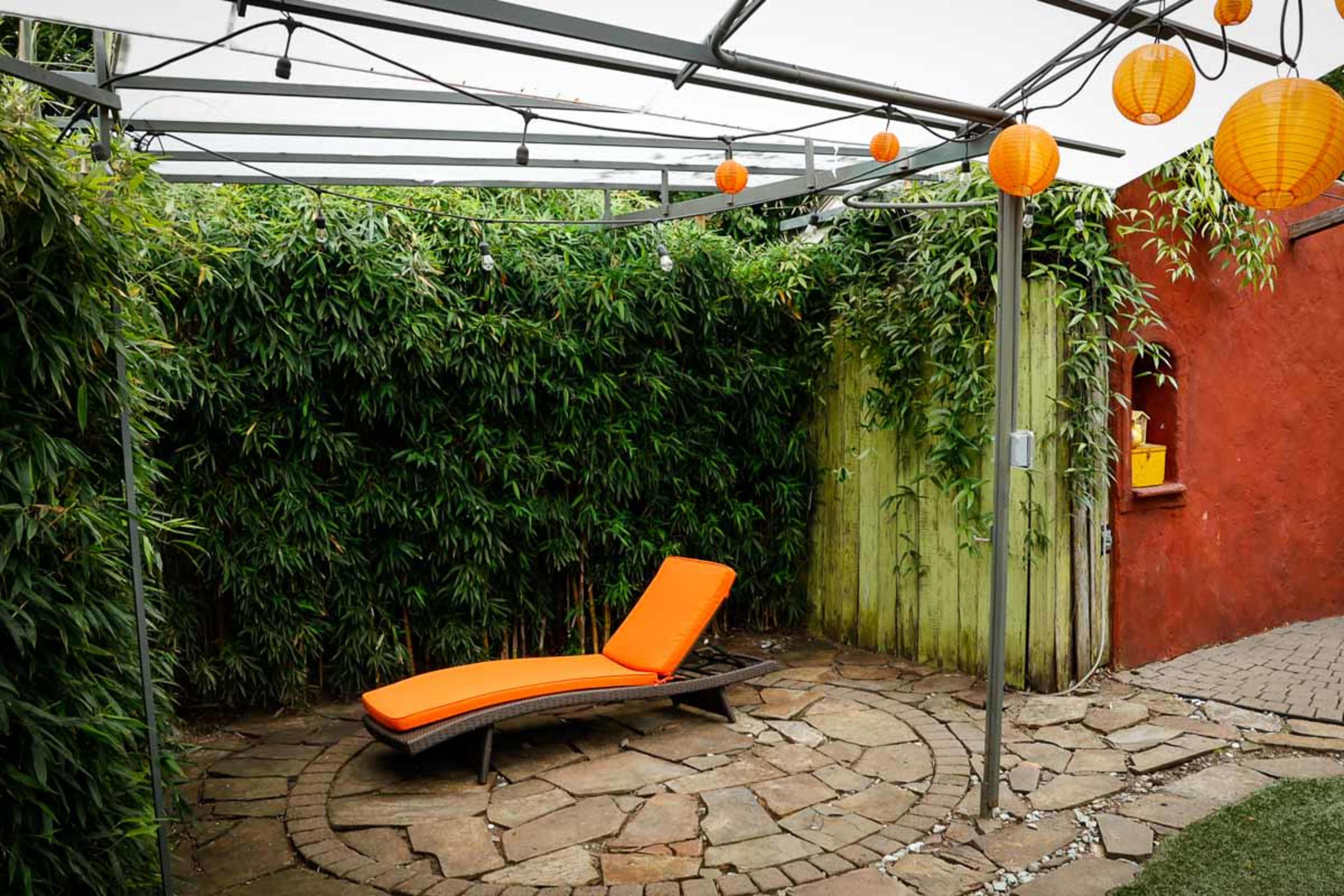 A lounge chair with an orange cushion is positioned under a covered area surrounded by bamboo and decorative orange lanterns.