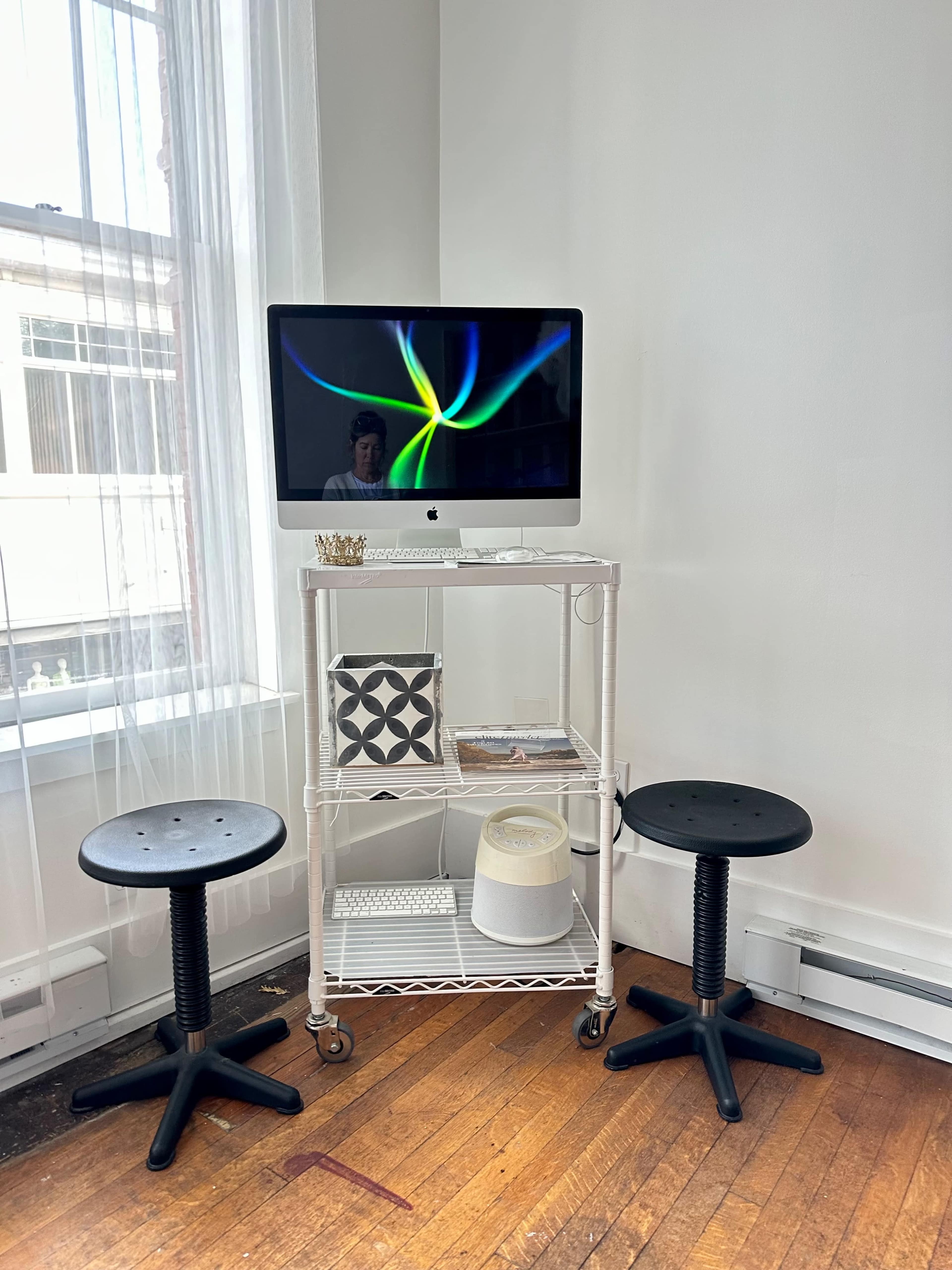 A metal cart holds an iMac computer and a few decorative items, flanked by two black stools in a well-lit room with large windows.