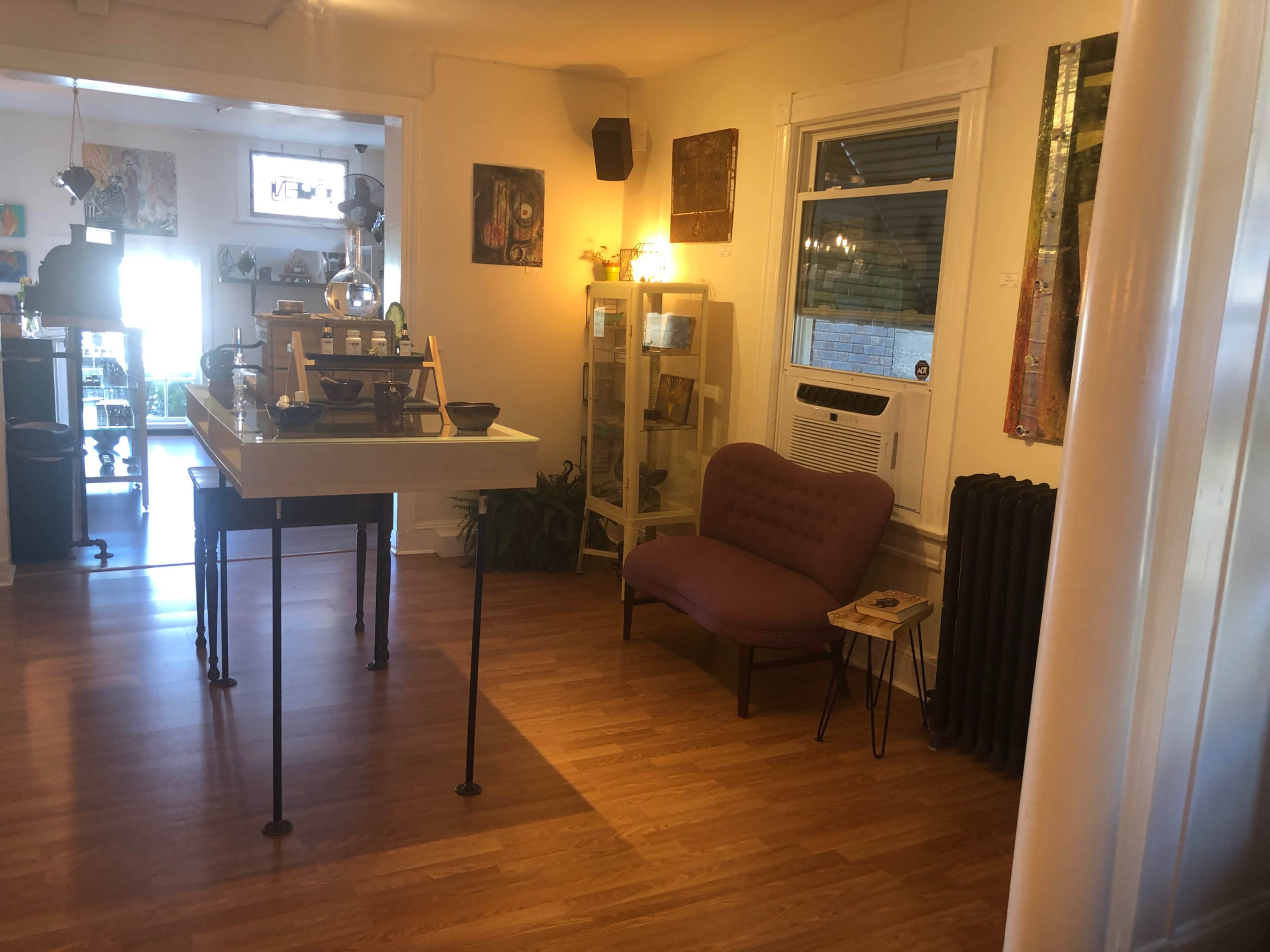 A cozy indoor space featuring a small table, a purple armchair, and a display cabinet, with wooden flooring and various artworks on the walls.