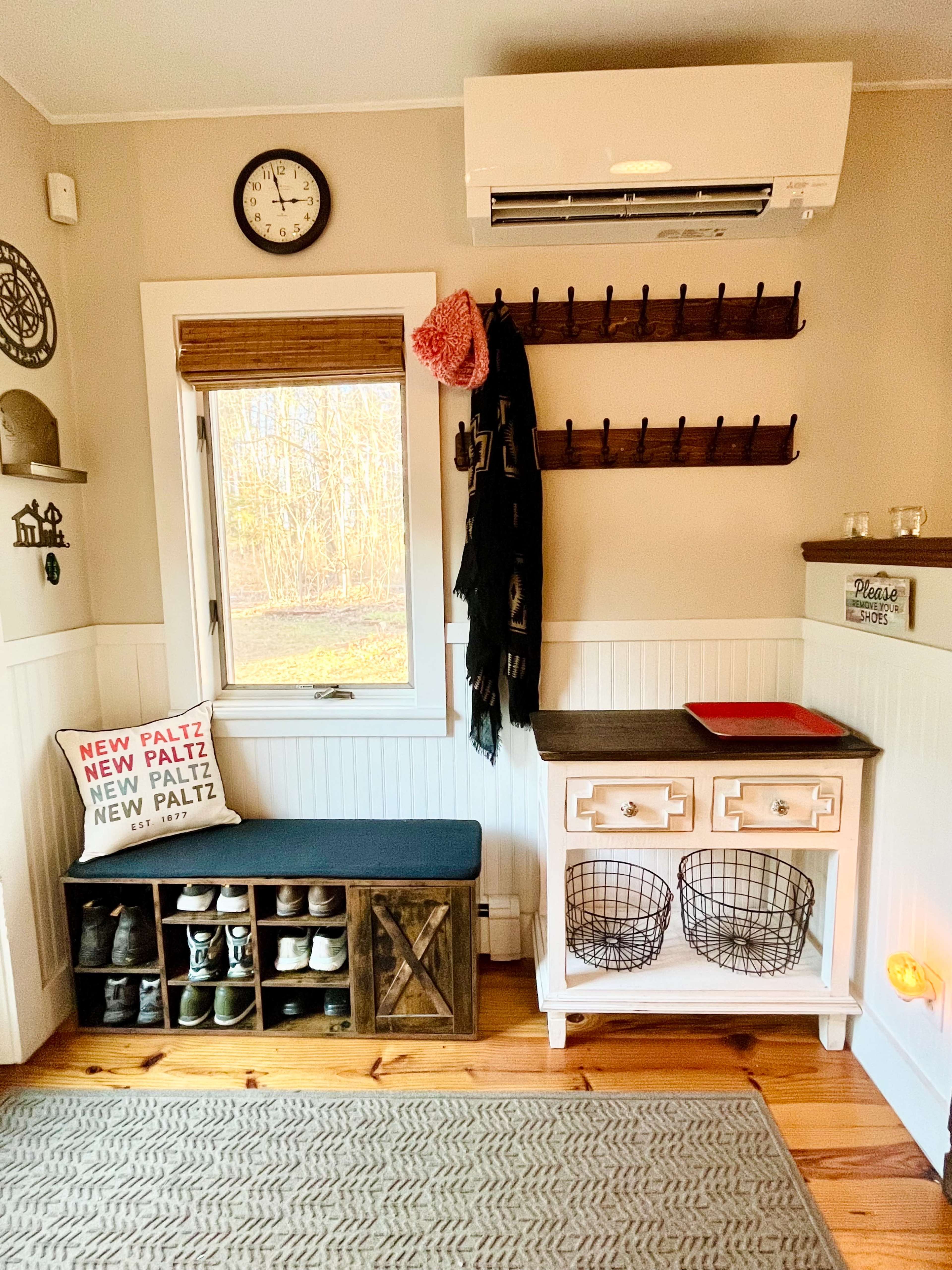 A cozy entryway featuring a shoe storage bench, a wall-mounted coat rack, a small table with decorative baskets, and a window with natural light illuminating the space.