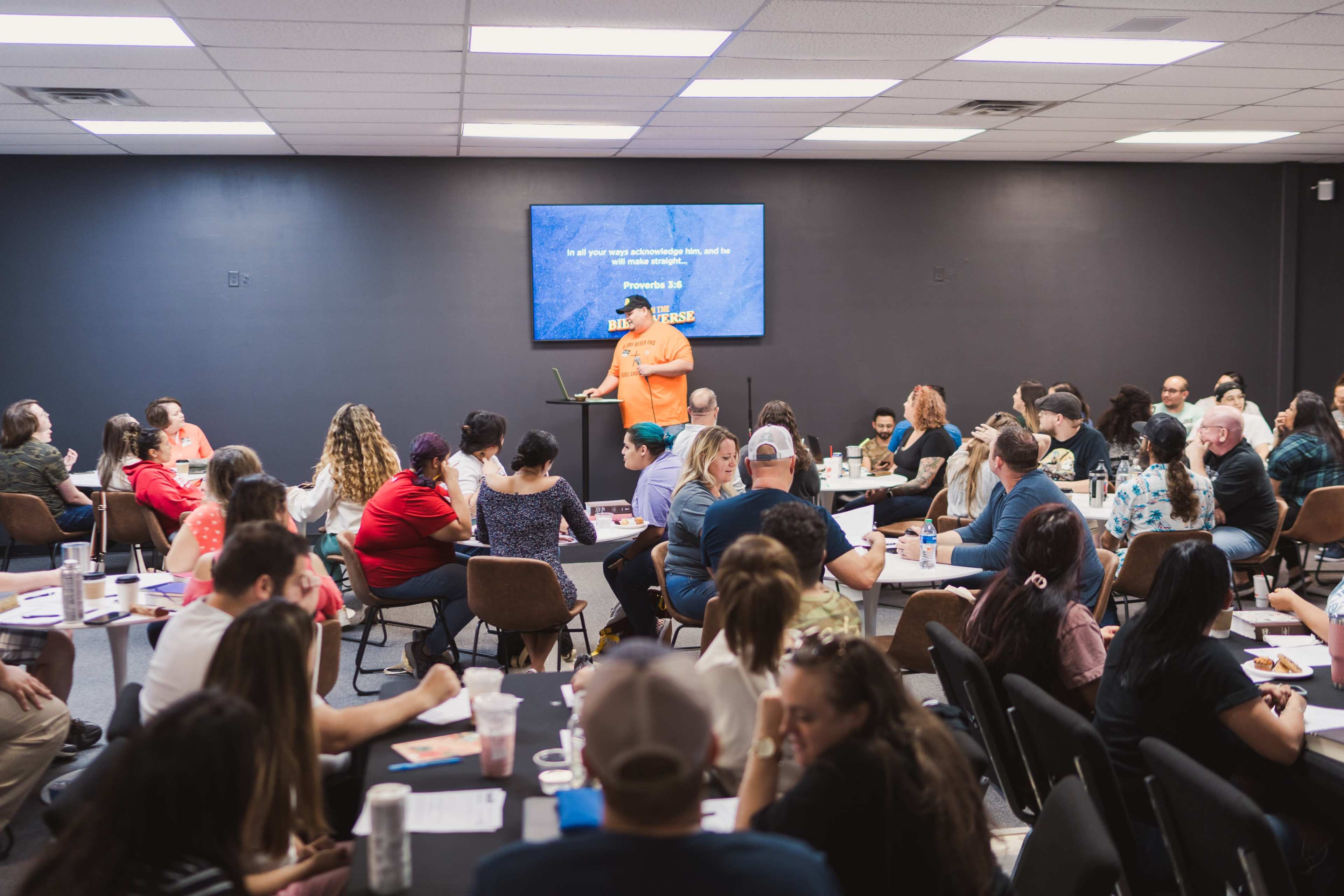 A large group of people sit at tables in a meeting room, attentively listening to a speaker in front of a screen displaying a presentation.