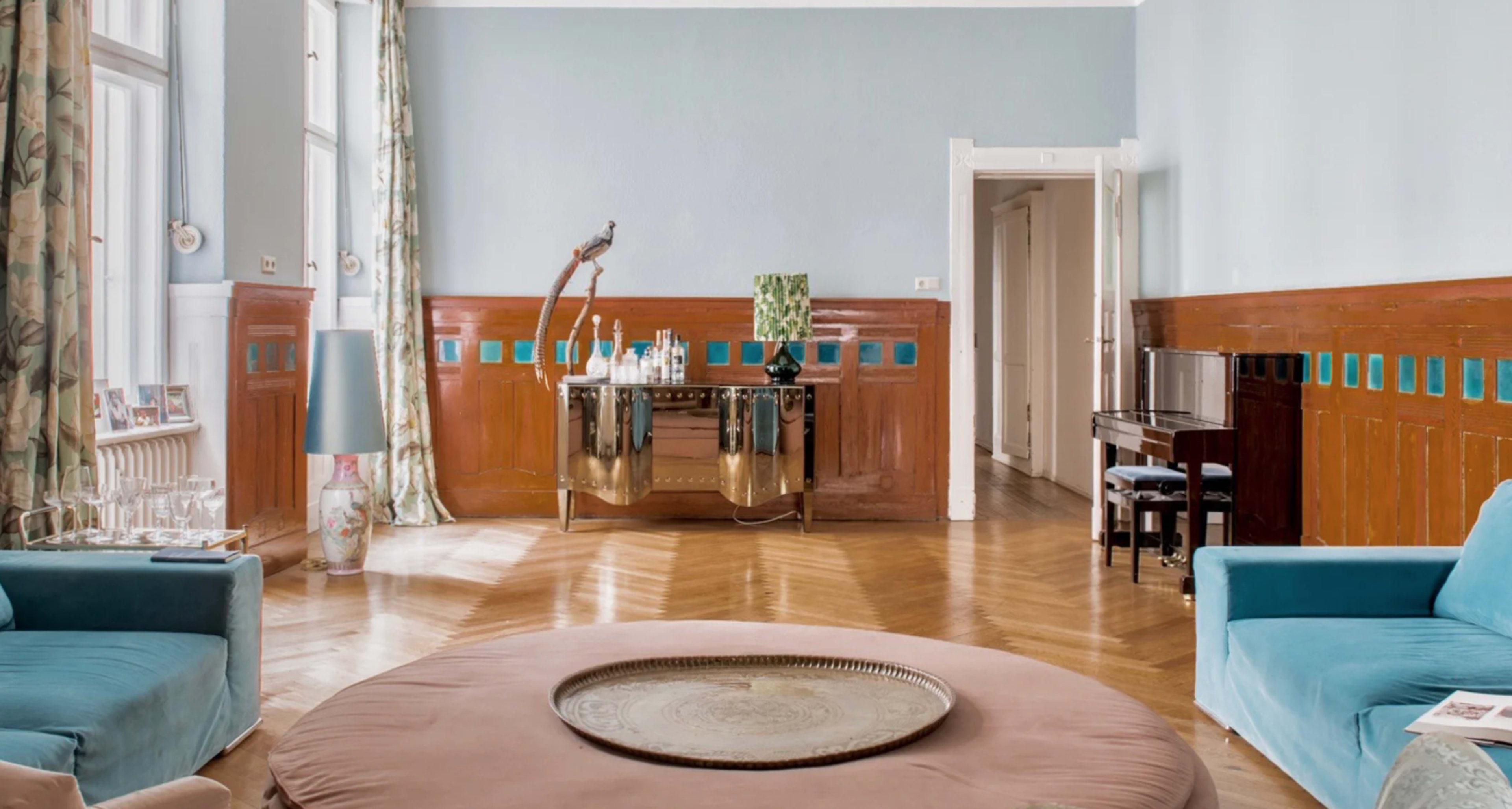 A light blue room with wooden paneling, featuring a round table in the center, a piano against one wall, and large windows with patterned curtains.