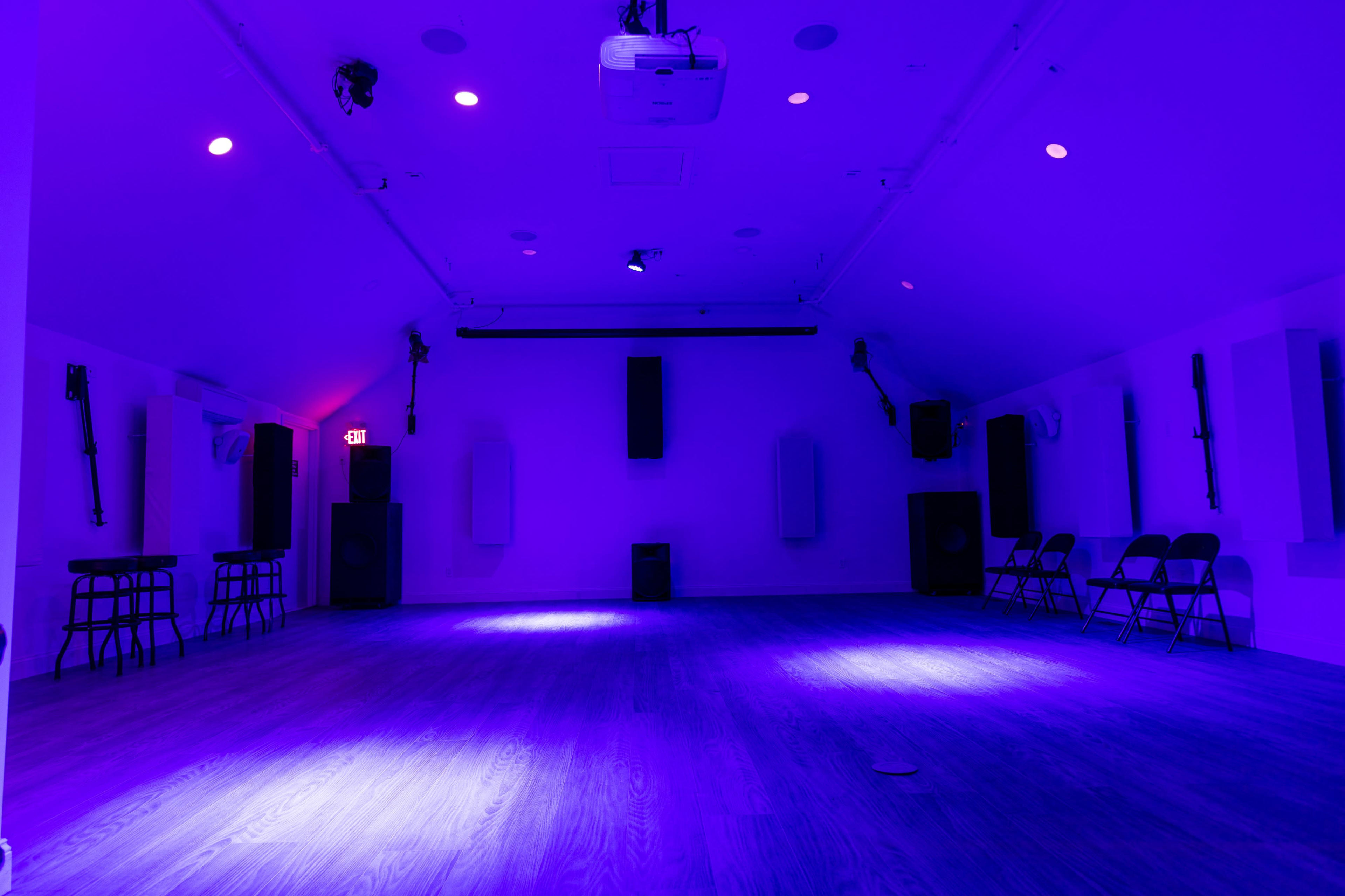 A spacious room with wooden flooring, lit by purple lights, featuring rows of chairs and audio equipment on the walls.