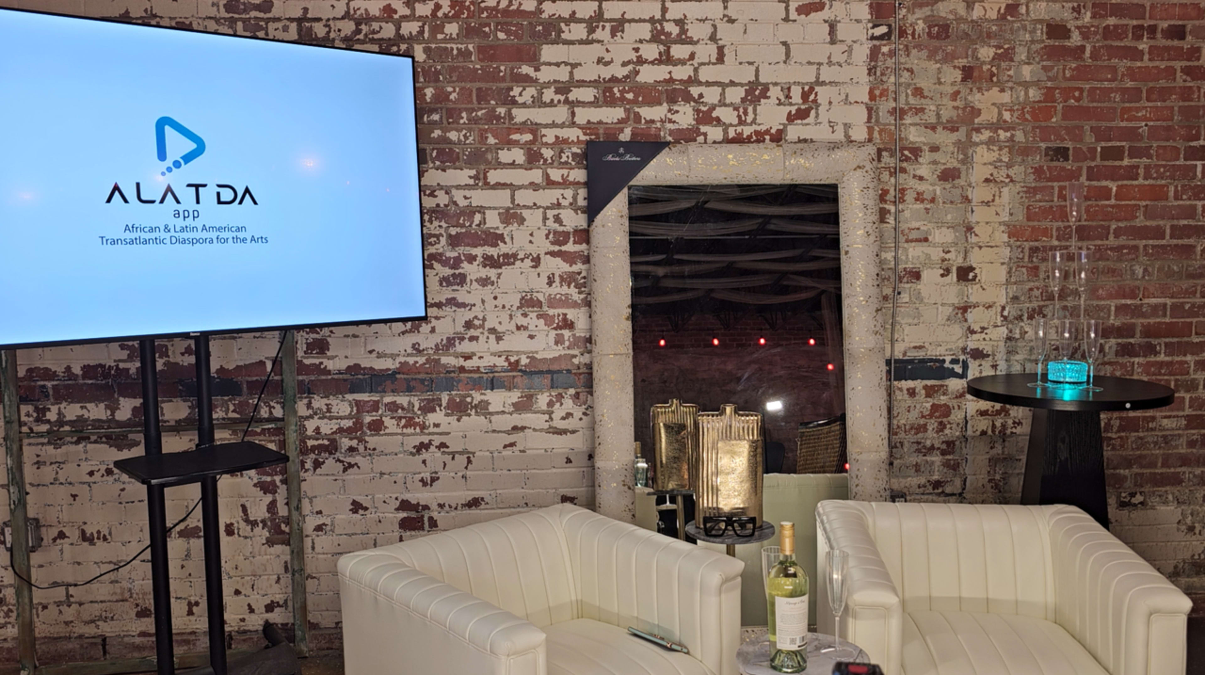 The image shows a modern event space featuring a large screen displaying the logo for ALATDA, a mirrored wall, and white leather chairs next to a small table with a bottle of wine.
