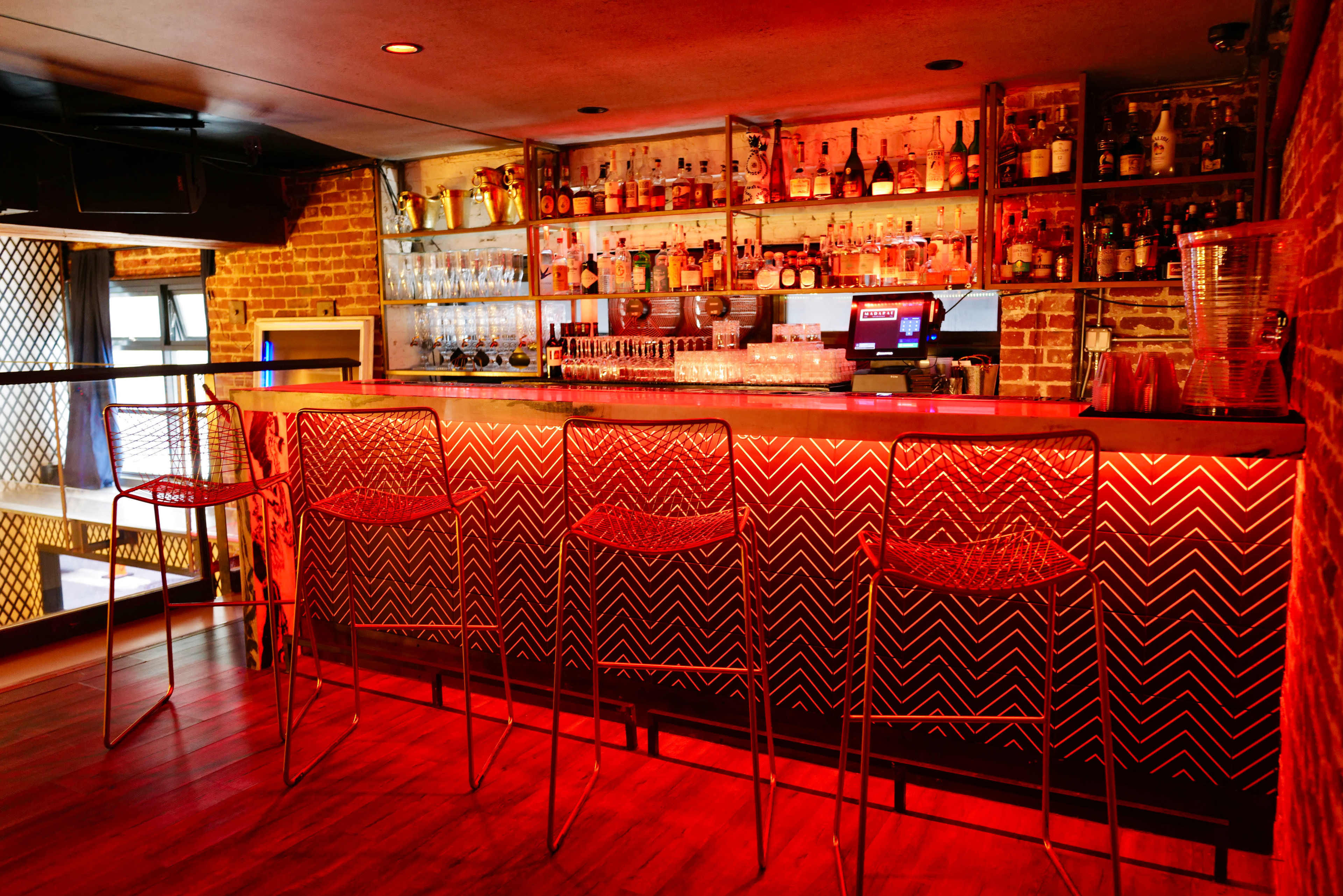 The image shows a well-lit bar area with a variety of bottles displayed on shelves behind the counter and four high chairs in front.