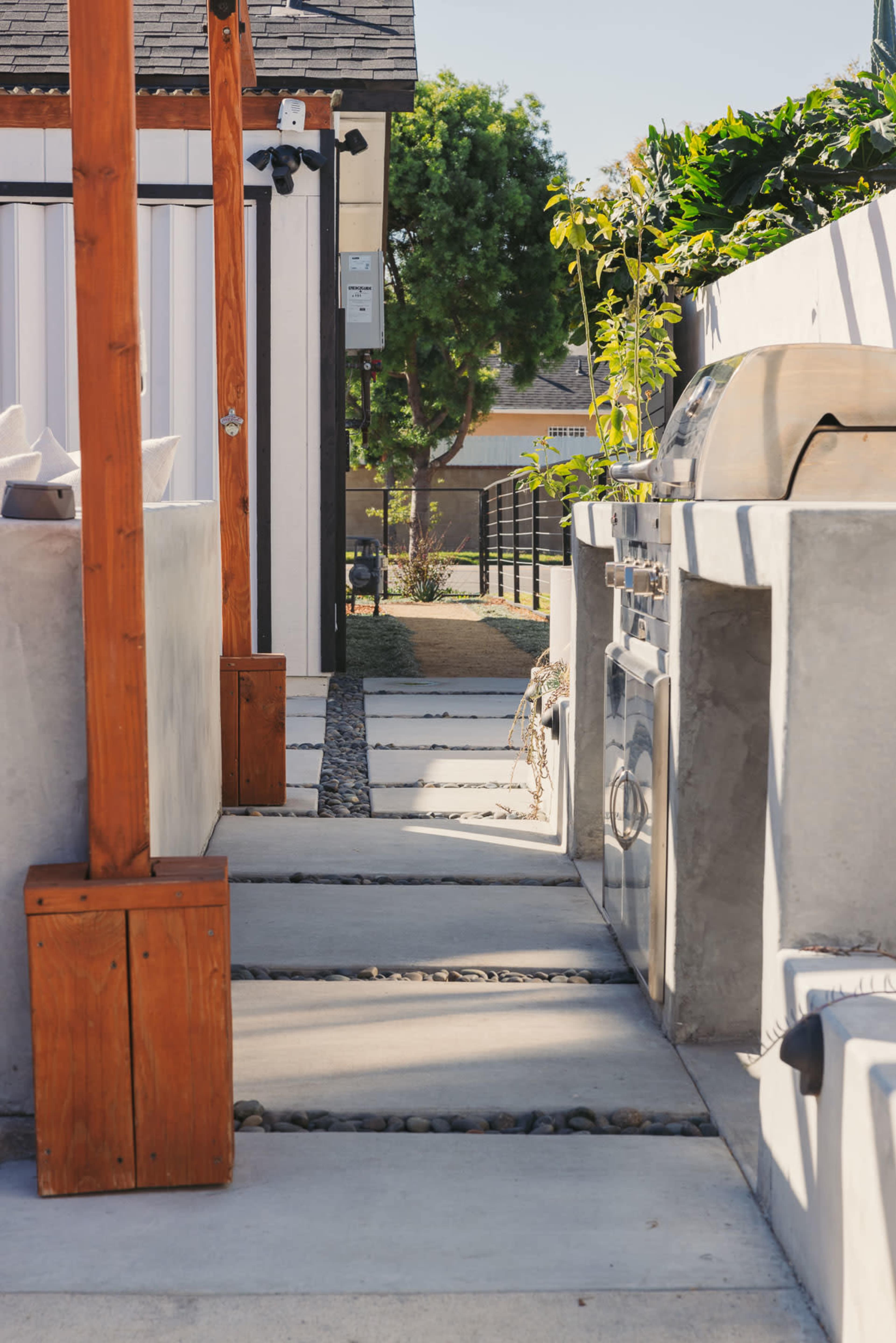 A narrow outdoor pathway made of concrete slabs, flanked by wooden posts and a stone garden area, leading to a built-in grill and an outdoor kitchen setup.