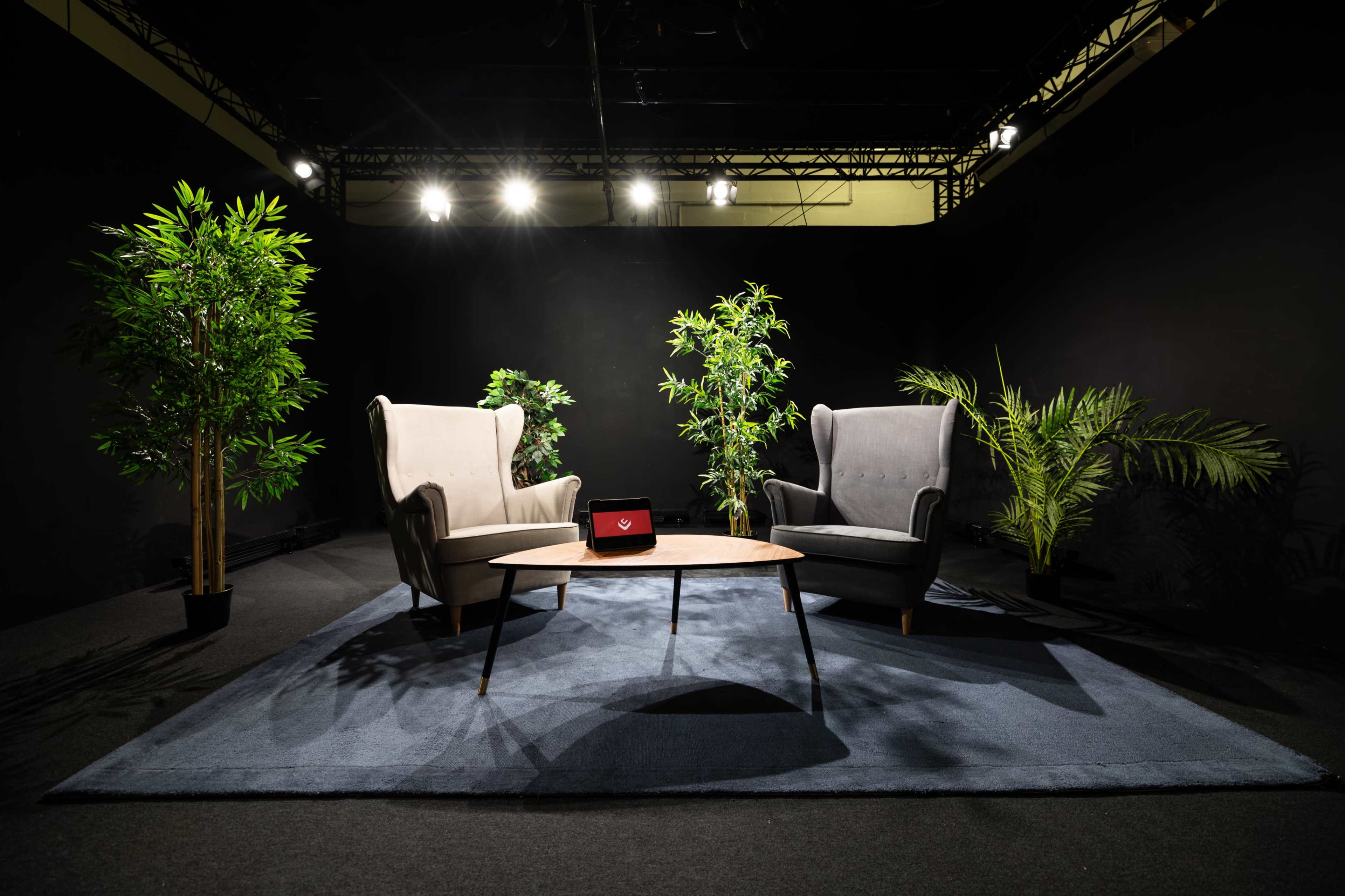 A cozy seating area features two armchairs facing each other, a round table in the center, and potted plants in a dimly lit space.
