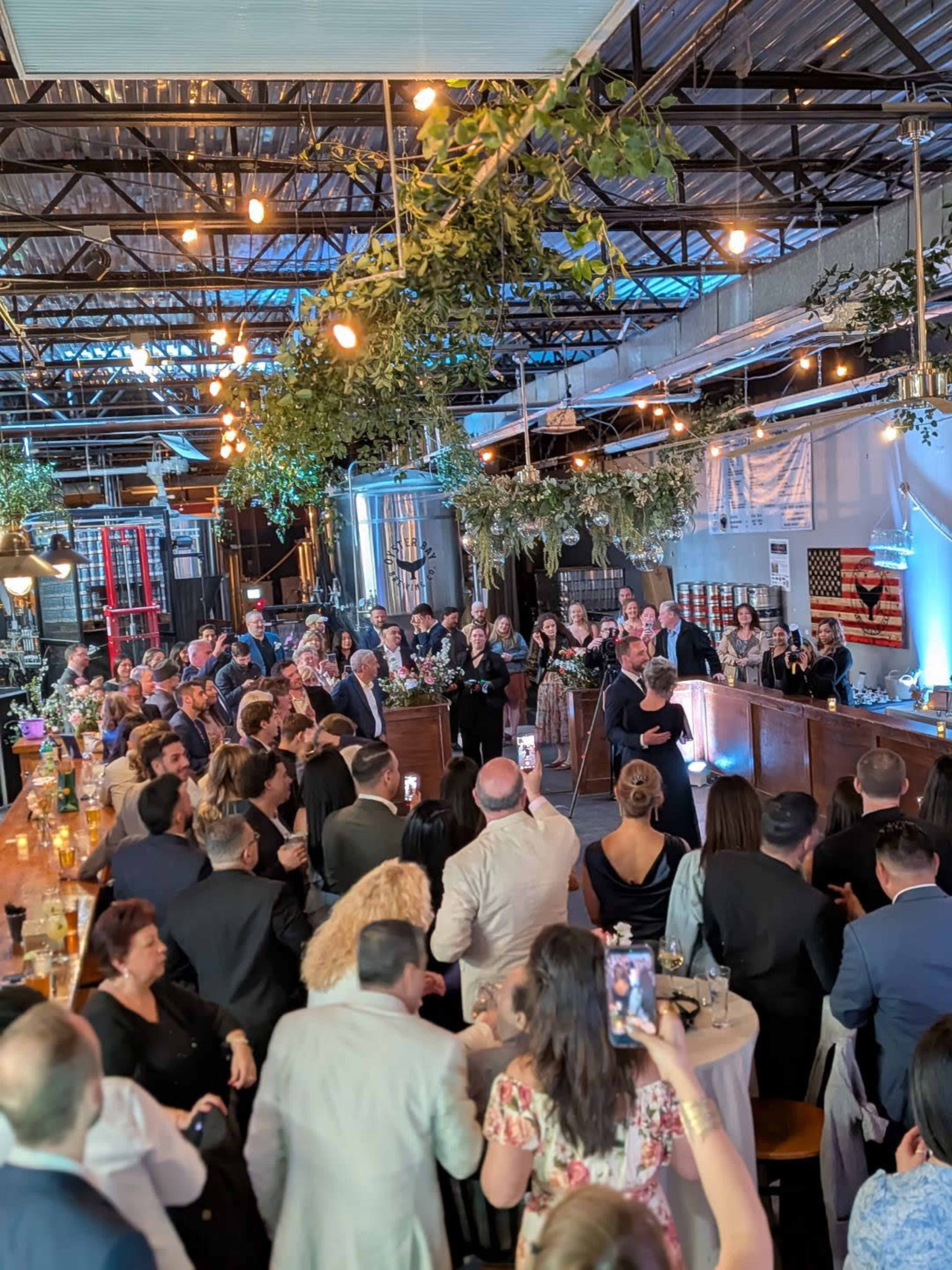 A large group of people gathers in a decorated industrial venue, with a bar at the front and various plants hanging from the ceiling.