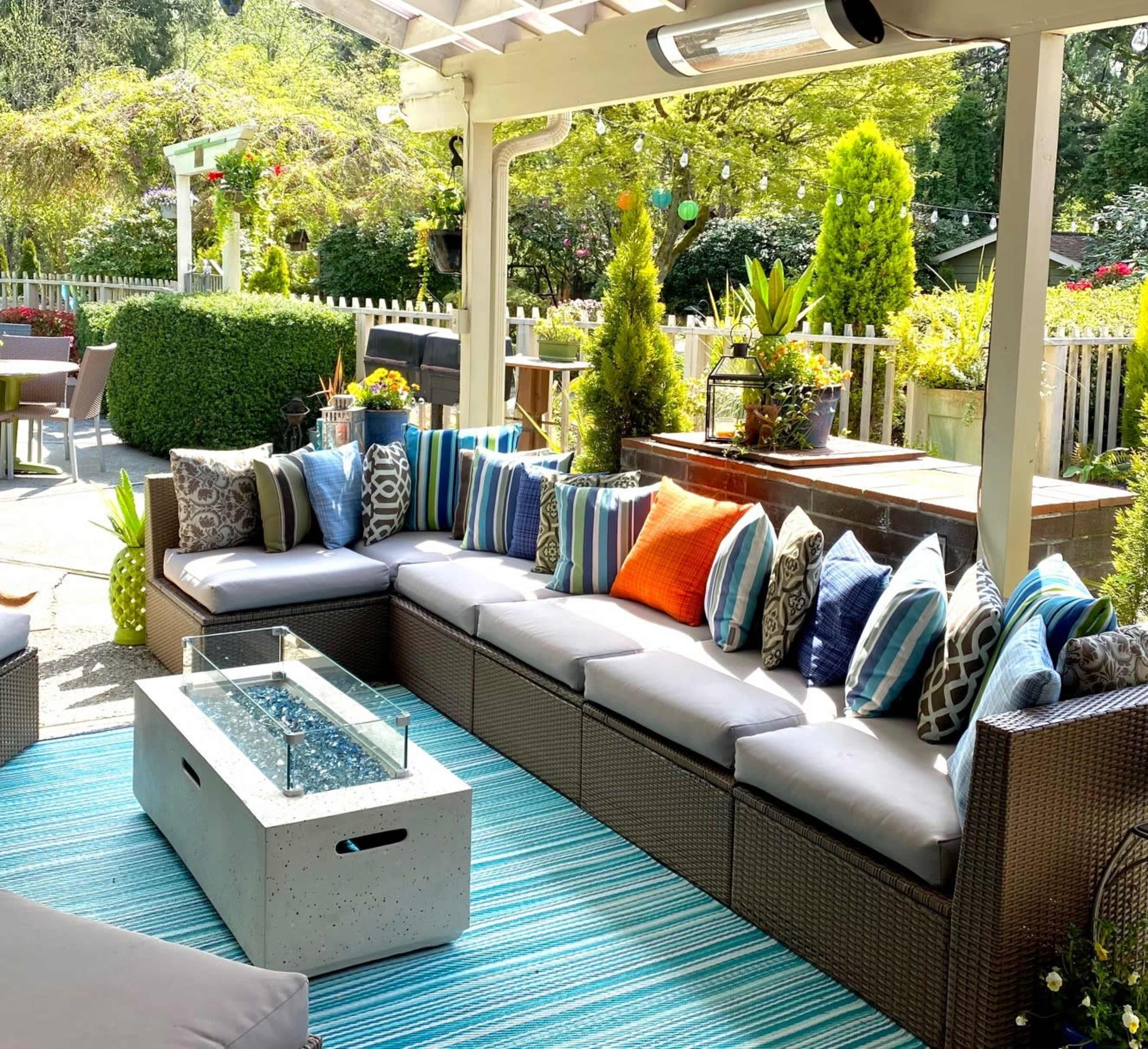 A patio area features a sectional sofa adorned with various throw pillows, a concrete fire table, and a striped outdoor rug, surrounded by greenery and decorative plants.