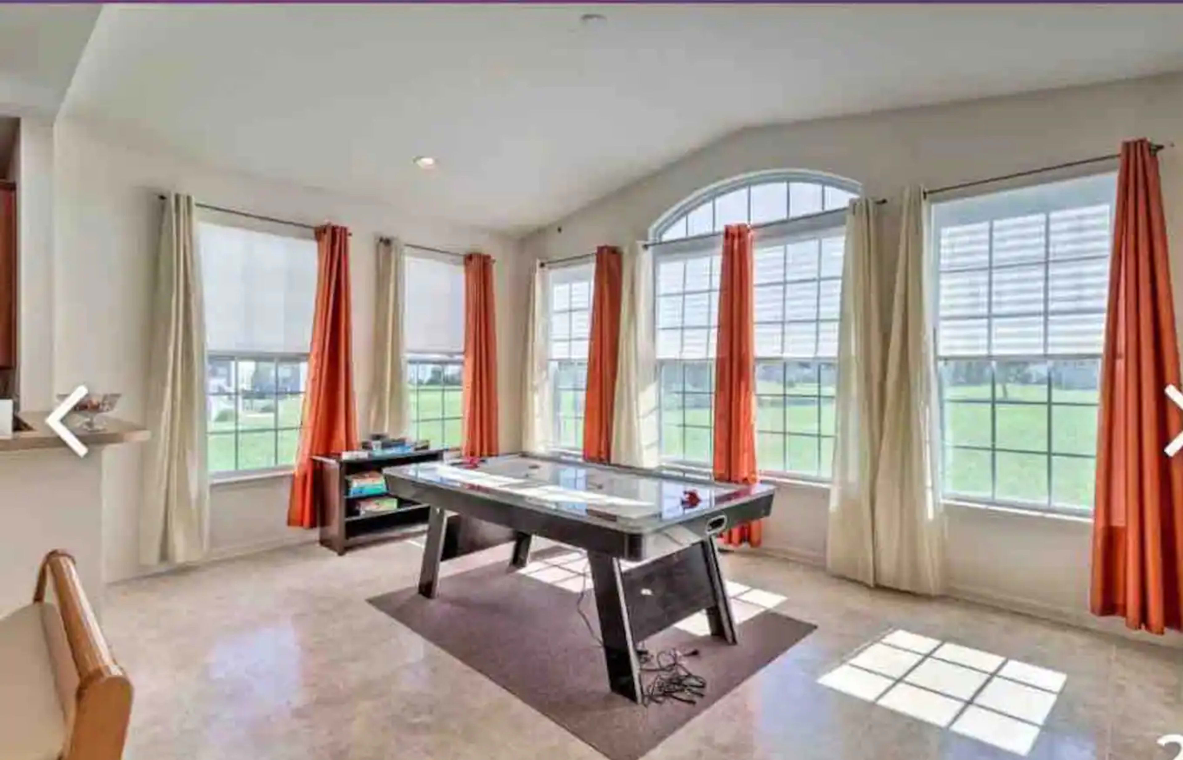 A bright room with large windows, featuring an air hockey table at its center and orange curtains hanging on the sides.