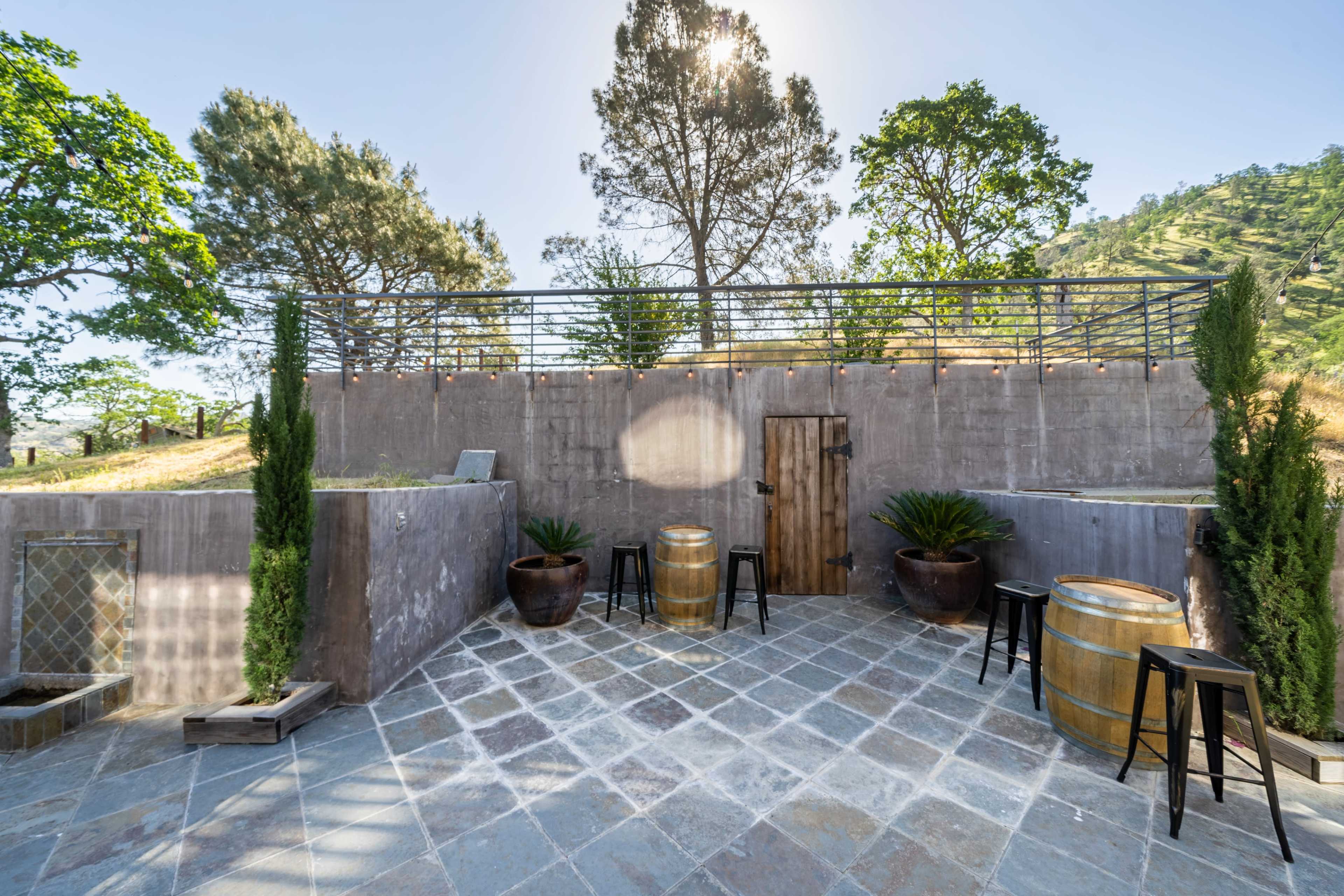 The image shows a spacious outdoor terrace with wooden barrels and tall plants, surrounded by trees and hills under a clear sky.