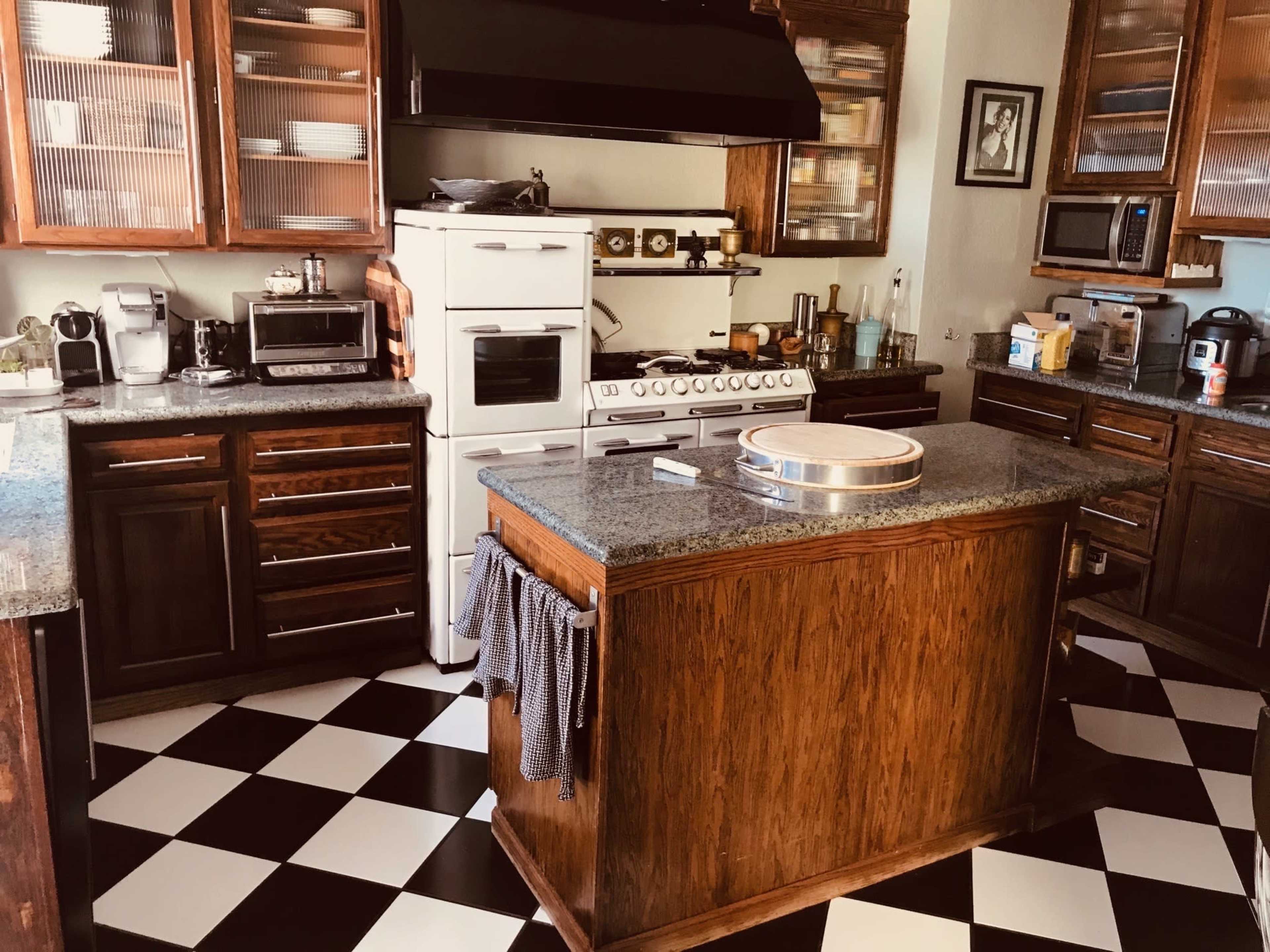 The image shows a kitchen featuring a central island with a granite countertop, wooden cabinets, and a checkered black-and-white floor.