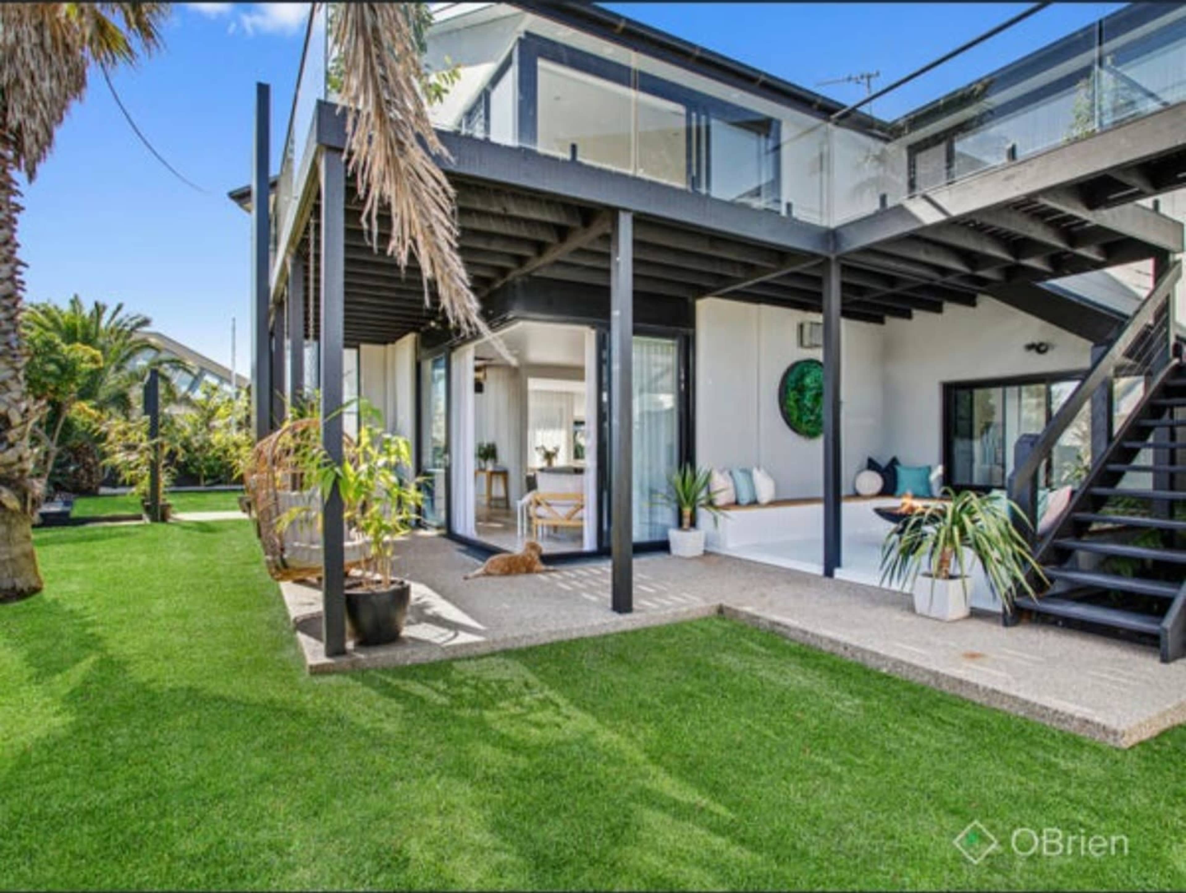 The image shows a modern two-story house with large windows, a terrace, and a well-maintained grassy yard surrounded by palm trees.