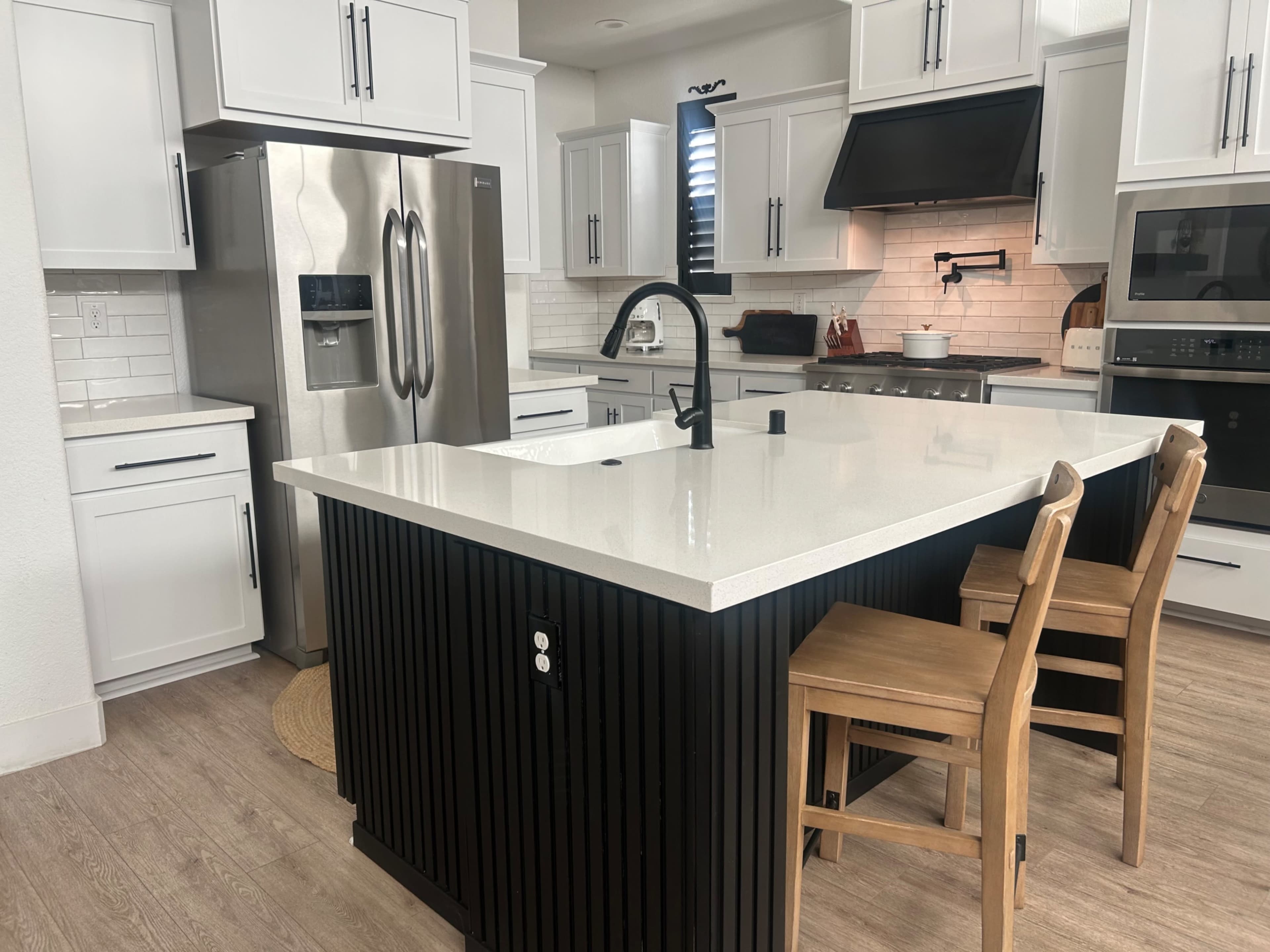 The image shows a modern kitchen with white cabinets, stainless steel appliances, a large island with a black finish, and two wooden bar stools.