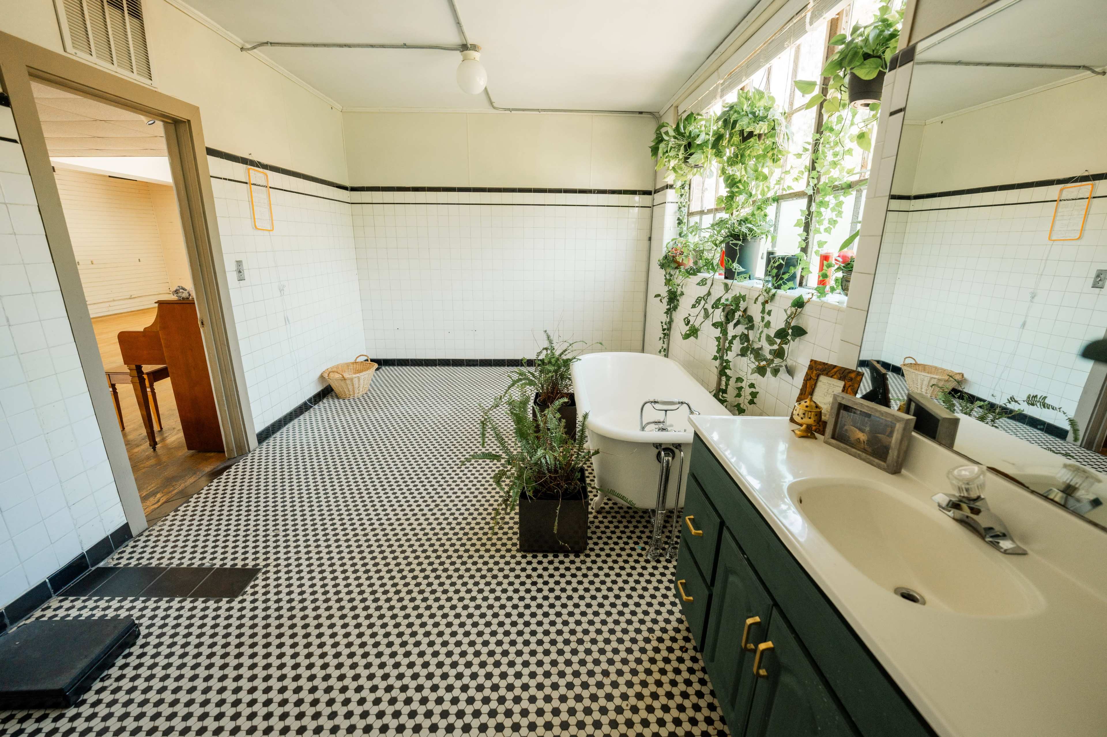 A bathroom with a vintage bathtub, a sink, and plants decorating the windowsill and floor, all set against black and white tiled walls and a checkered floor.