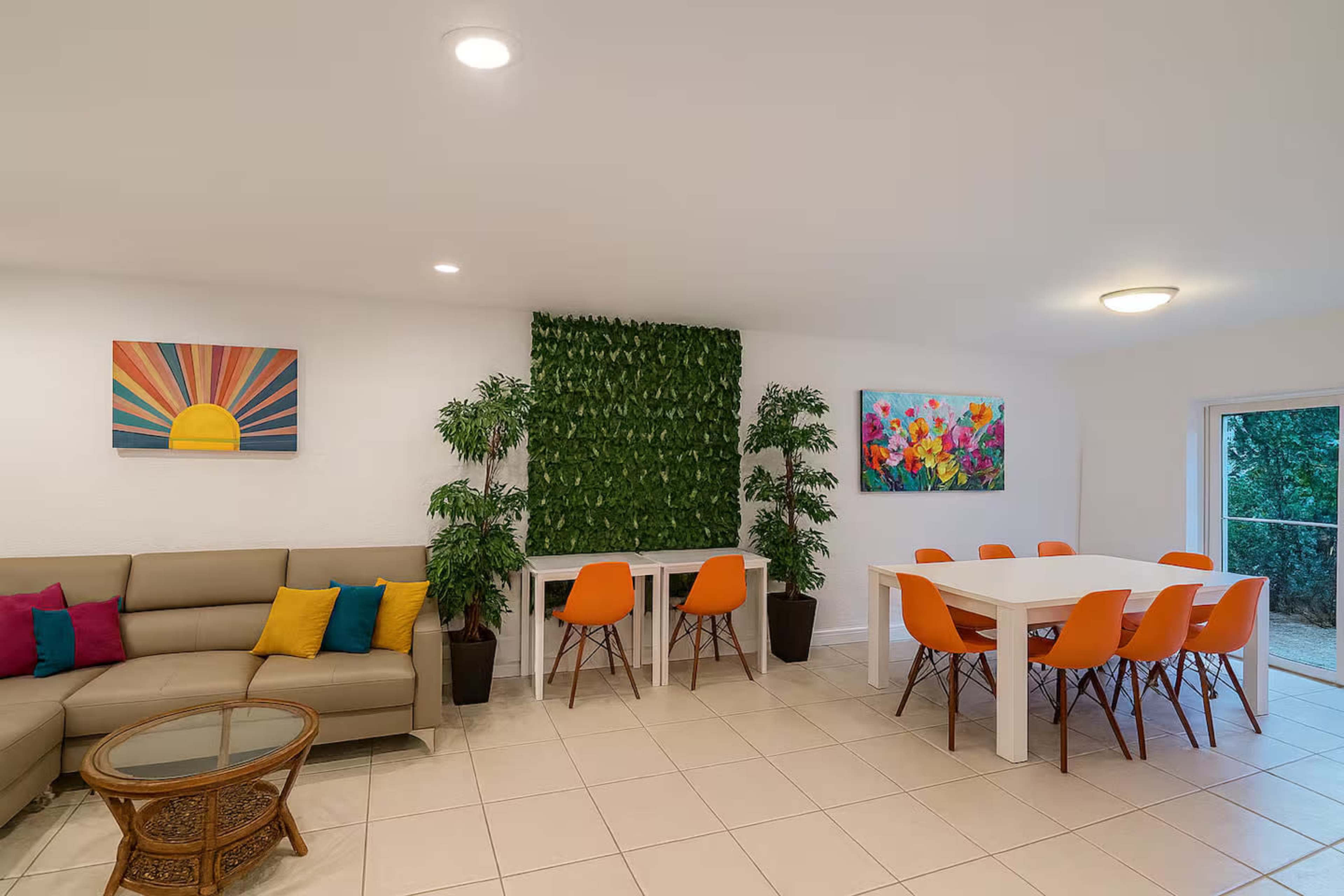 The image shows a bright, modern living space featuring a beige sofa with colorful cushions, a dining table with orange chairs, and wall art, along with greenery on one wall.