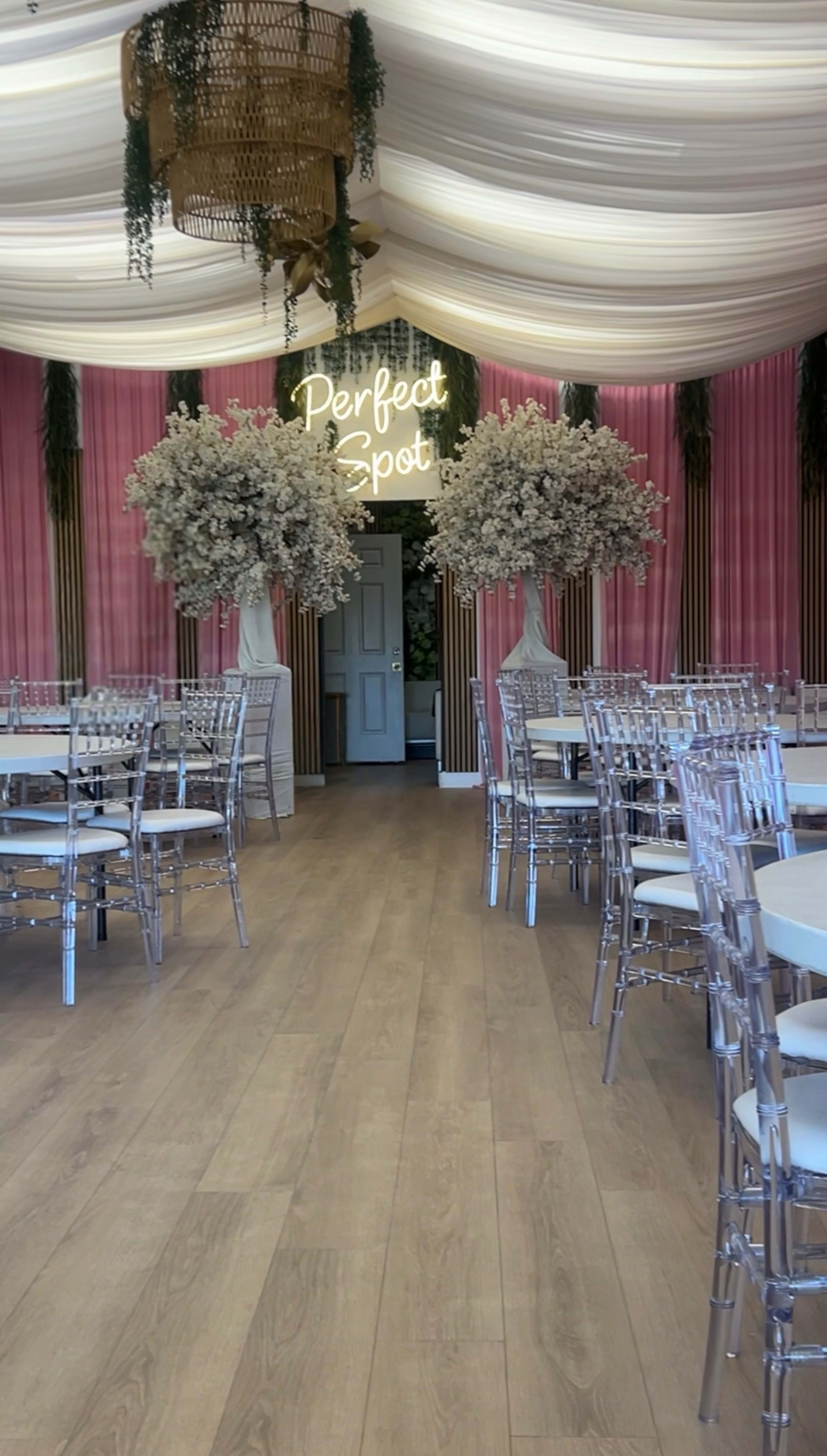 The image shows an elegantly decorated interior with large floral arrangements, round white tables, and transparent chairs under a draped ceiling, featuring a neon sign that reads "Perfect Spot."