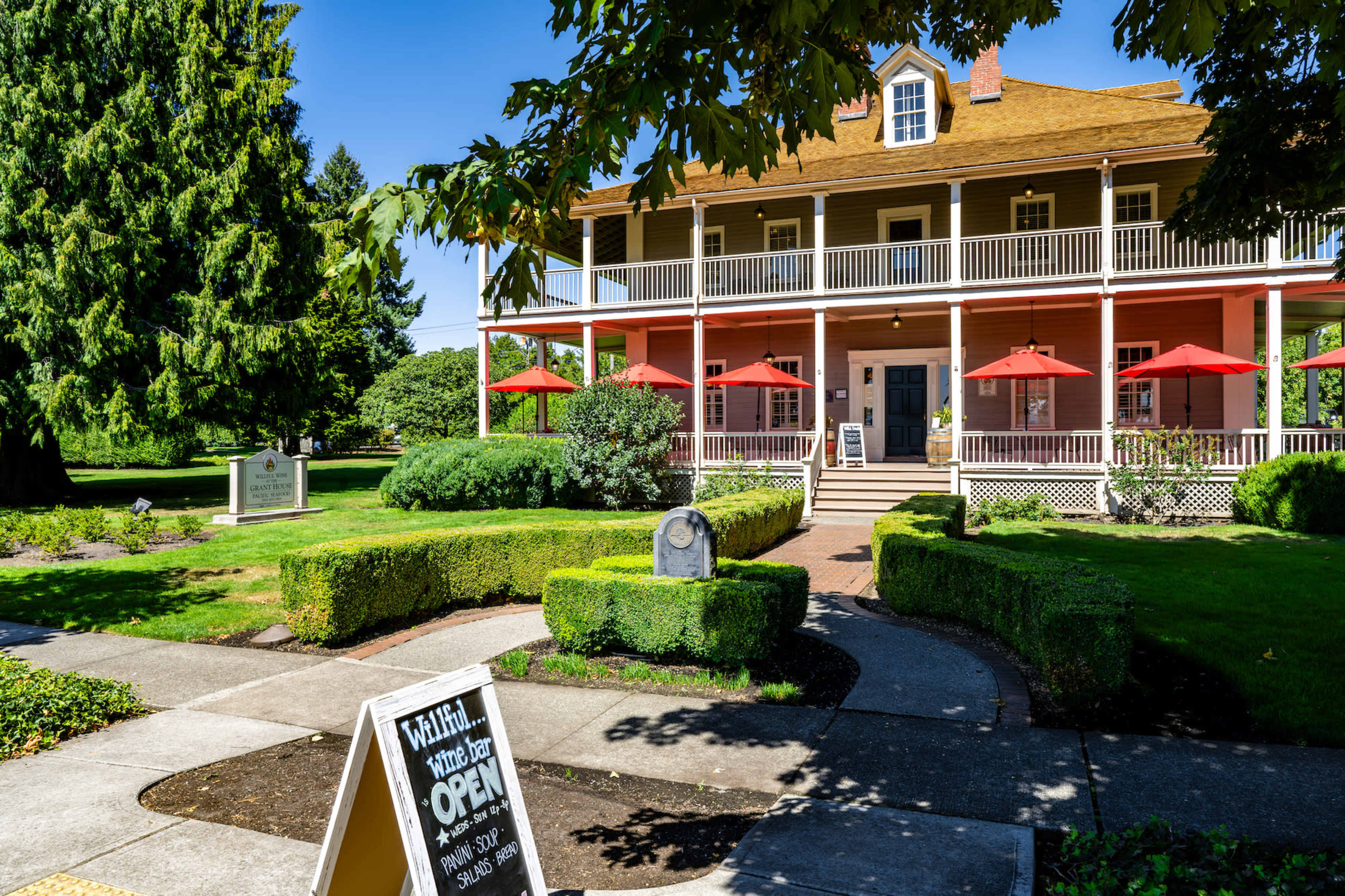A two-story pink building with a large porch and red umbrellas is surrounded by well-maintained greenery and shrubs.
