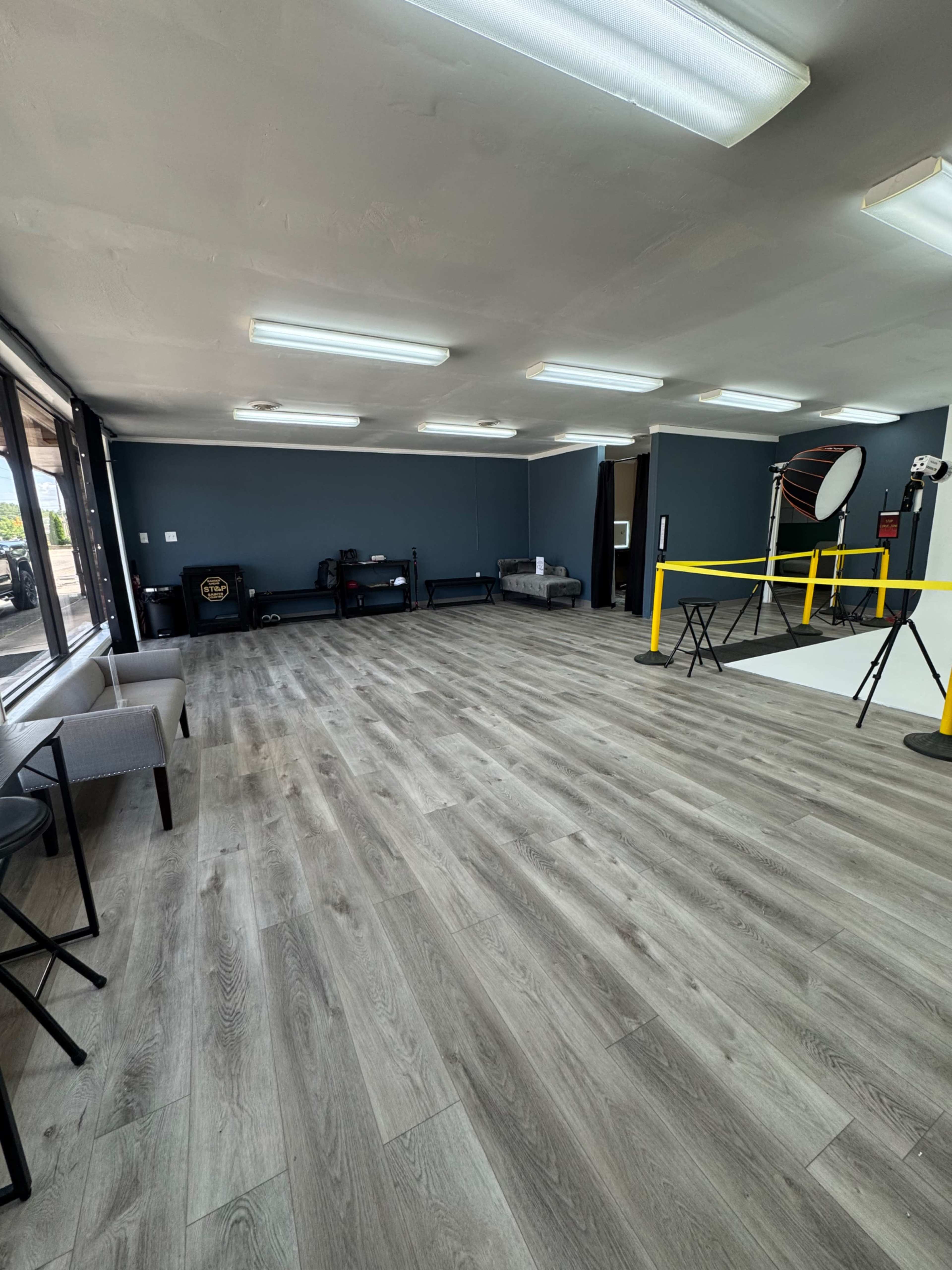 The image shows an empty studio space with gray walls, wooden flooring, and various photography equipment set up against one side.